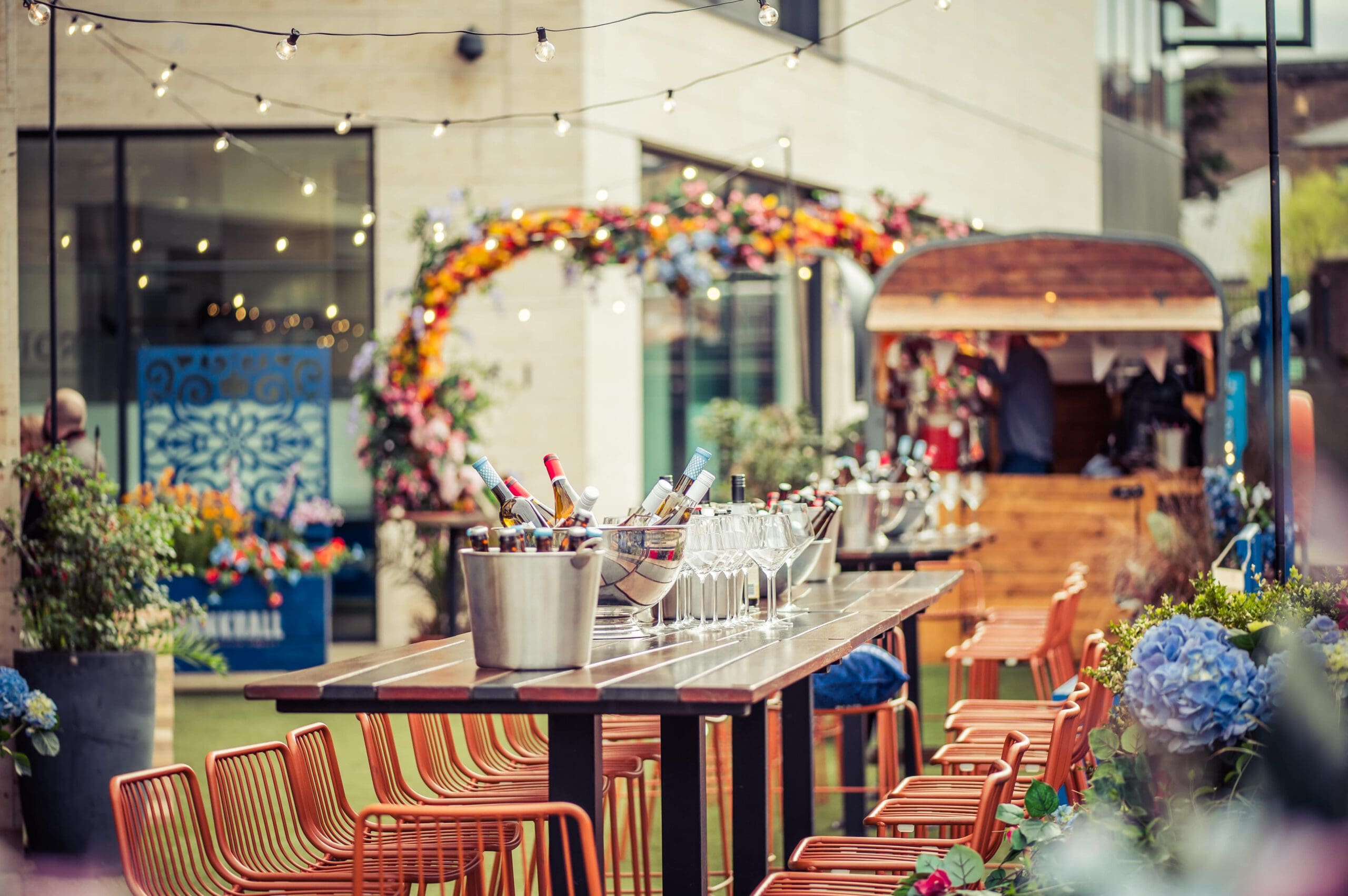 Outdoor event set-up with a long table lined with orange chairs, buckets of ice and wine bottles, and glasses. Fairy lights hang above, and a colourful floral arch is in the background near a wooden stall.