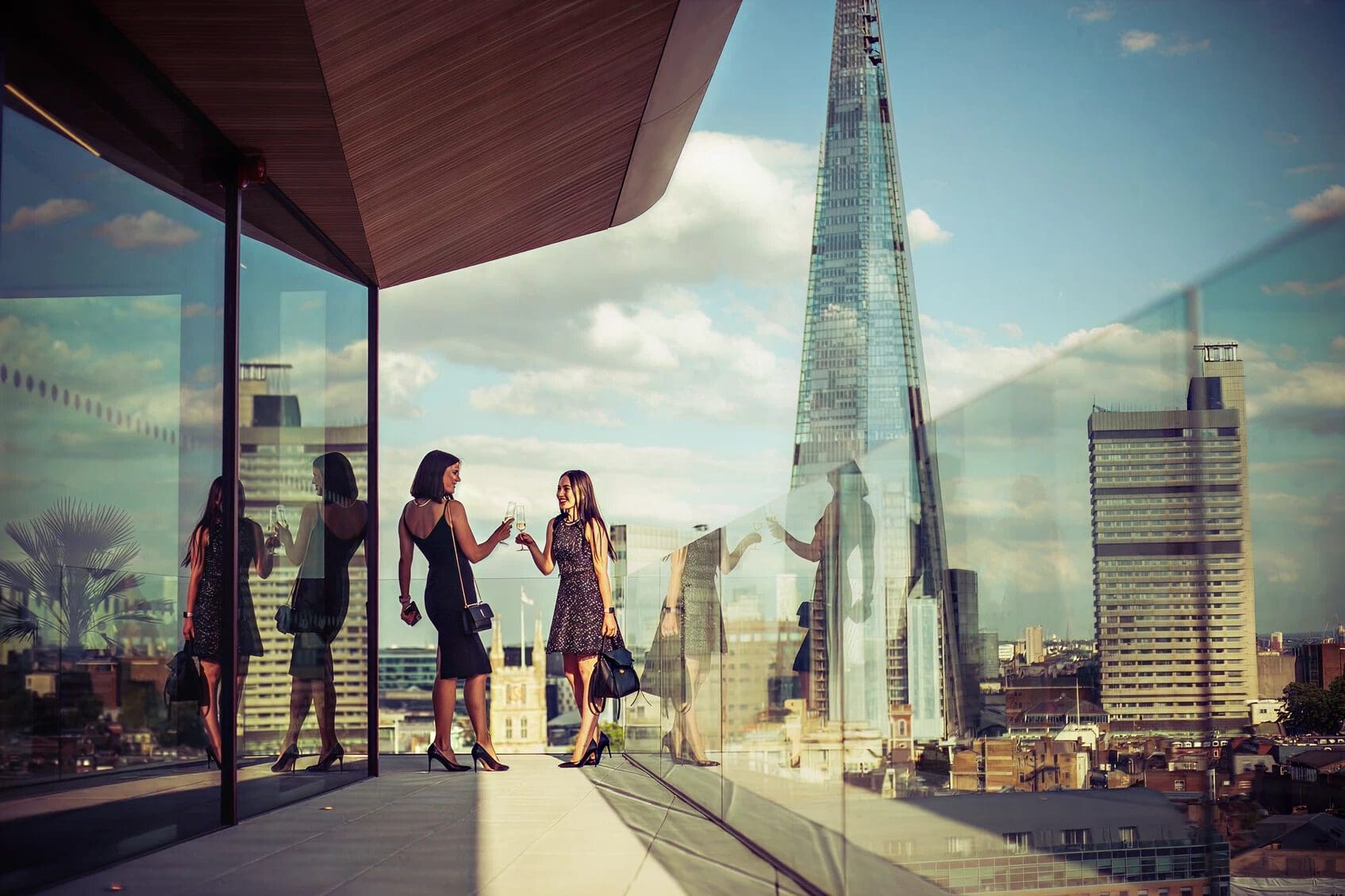 Two women clink glasses on a modern rooftop terrace with glass balustrades, overlooking a cityscape featuring the Shard skyscraper on a sunny day.