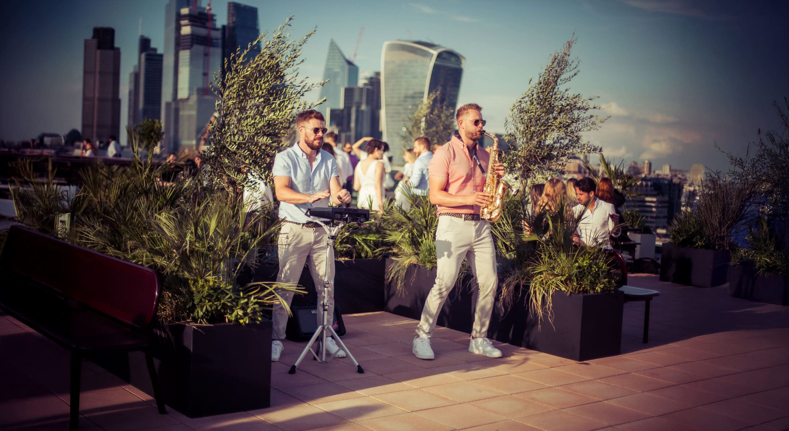Two men play music on a rooftop, one with a saxophone and one with an electronic drum pad, surrounded by plants. People are mingling in the background with a city skyline beyond.