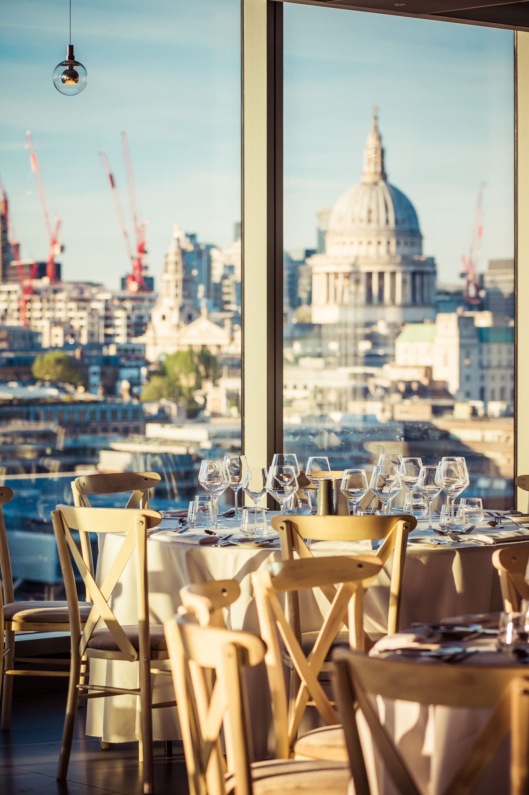 A sunlit restaurant with elegantly set tables and wooden chairs, featuring large windows offering a cityscape view, including St Paul's Cathedral and construction cranes in the background.