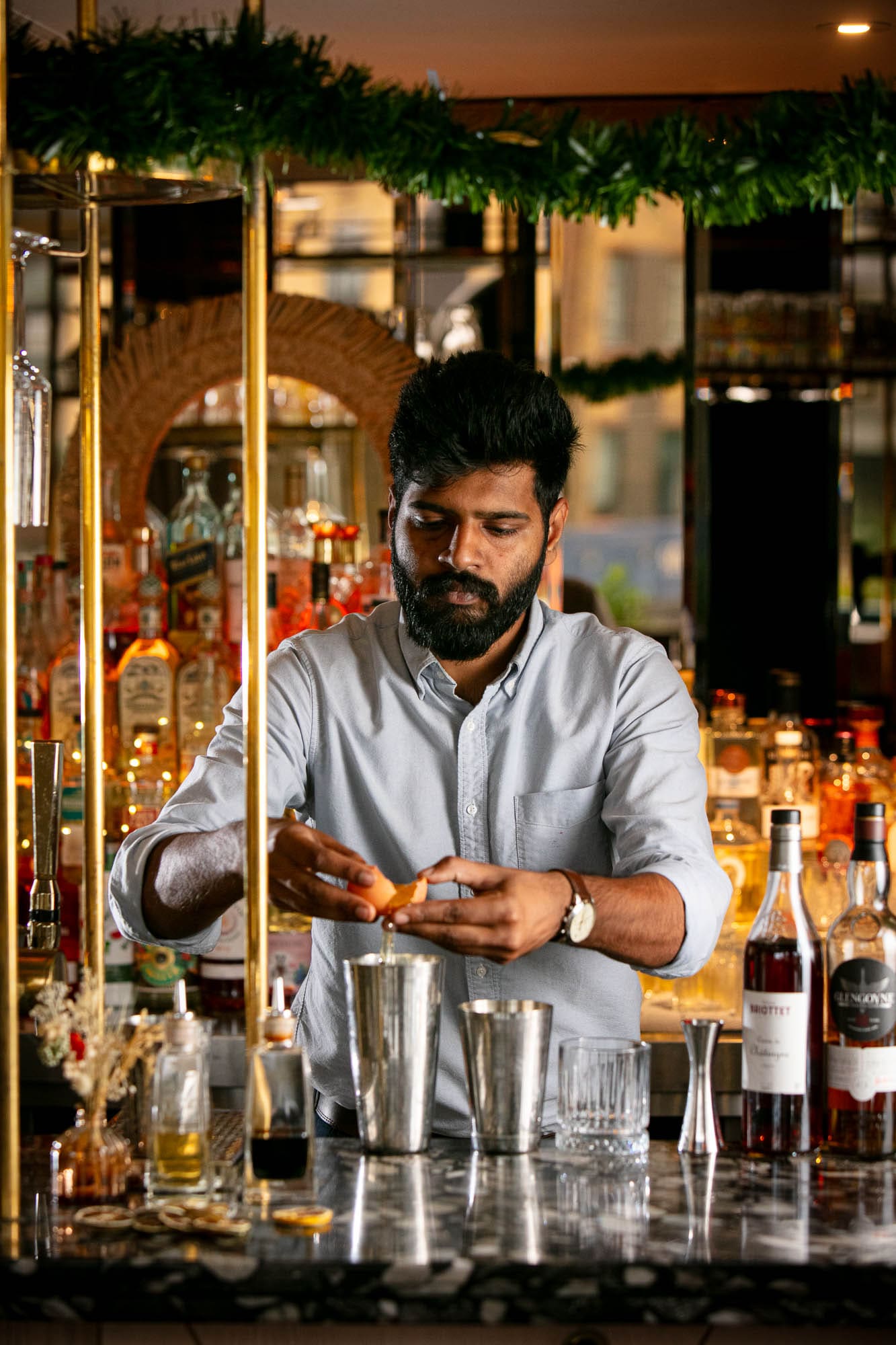 A barman with a beard, wearing a light blue shirt, prepares a cocktail behind a bar counter with bottles and glasses, breaking an egg over a shaker in a warmly lit bar setting.