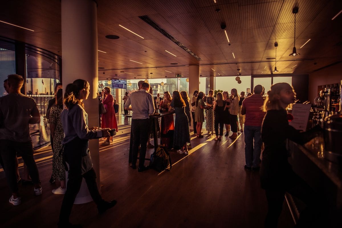 A group of people socialises in a modern, sunlit indoor space with wooden floors and large windows, casting long shadows. Some stand at a counter, while others mingle in small groups.