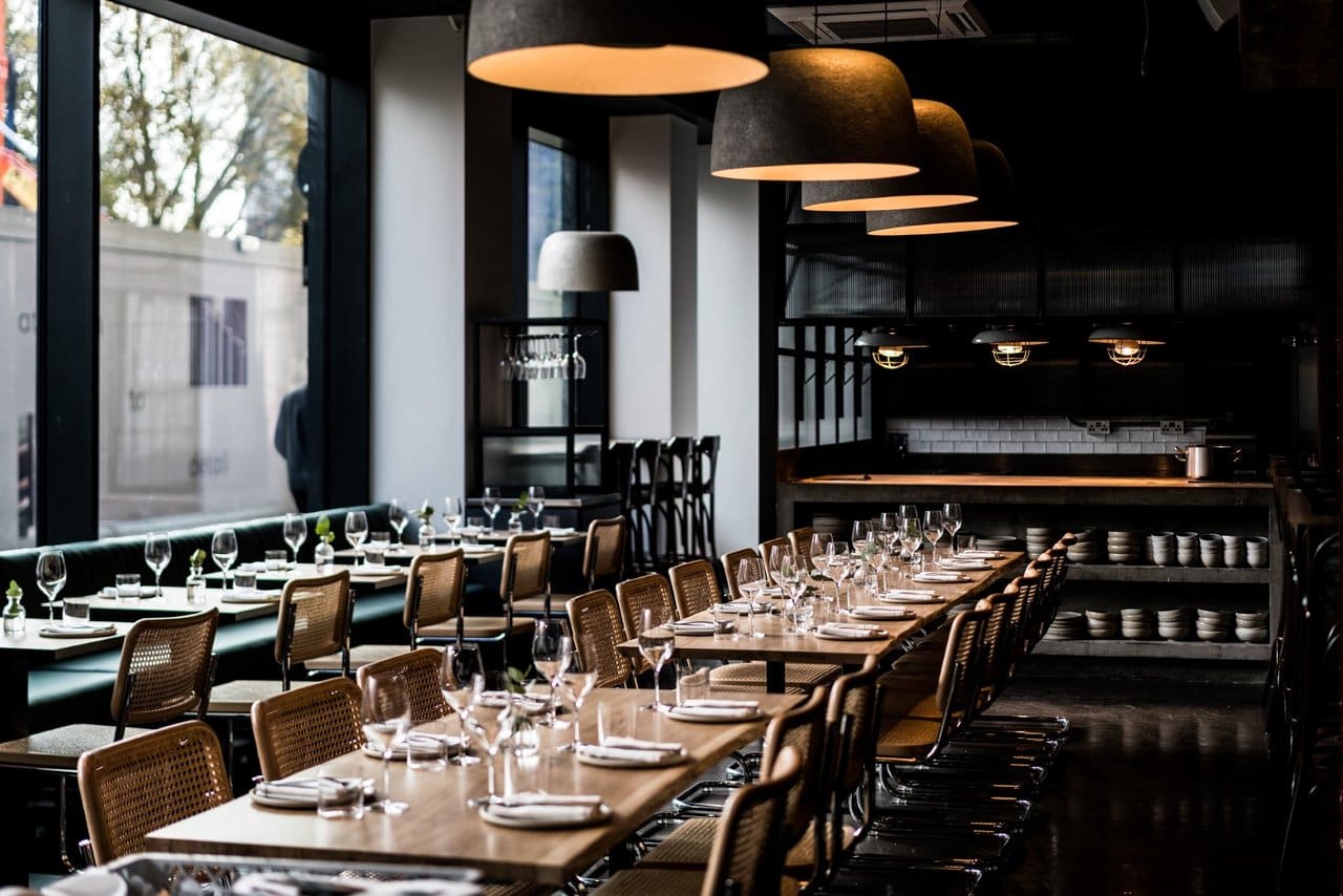 A modern restaurant with long wooden tables set with glasses, plates, and cutlery. Woven chairs line the tables, and large pendant lights hang from the ceiling. Natural light enters through large windows on the left.