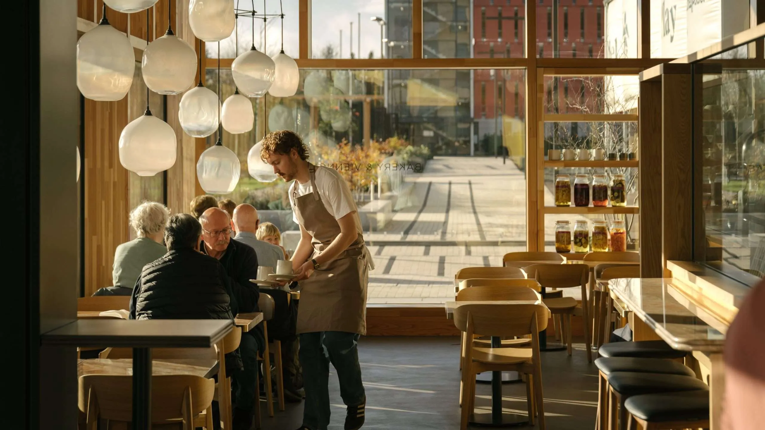 A waiter wearing an apron brings drinks to three people seated at a table inside a sunlit café with large windows, wooden furniture, hanging globe lights, and a view of an outdoor terrace.