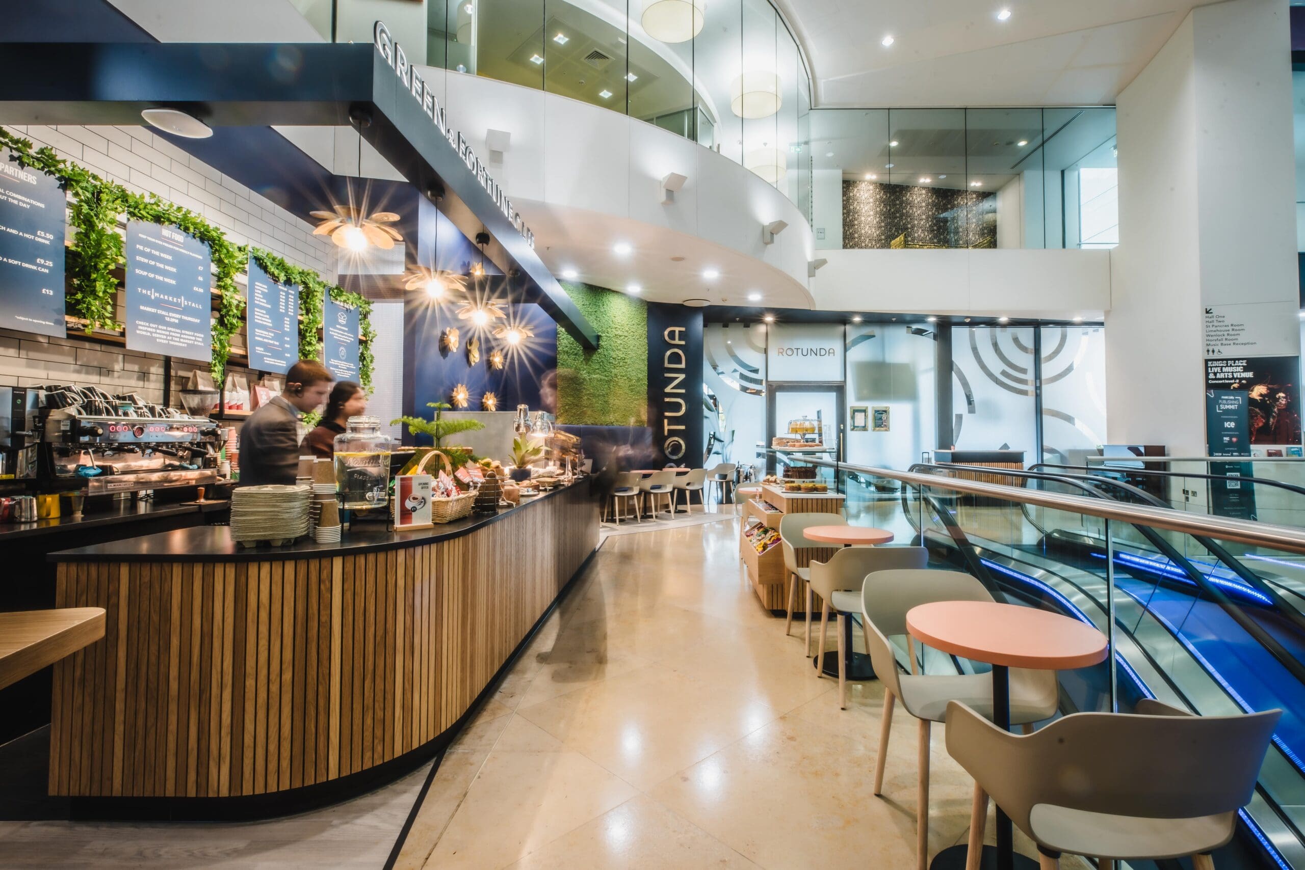 Modern café with curved wooden counter, two staff preparing drinks, menu boards on the wall, indoor plants, small tables and chairs, and large windows letting in natural light. The space appears bright and inviting.