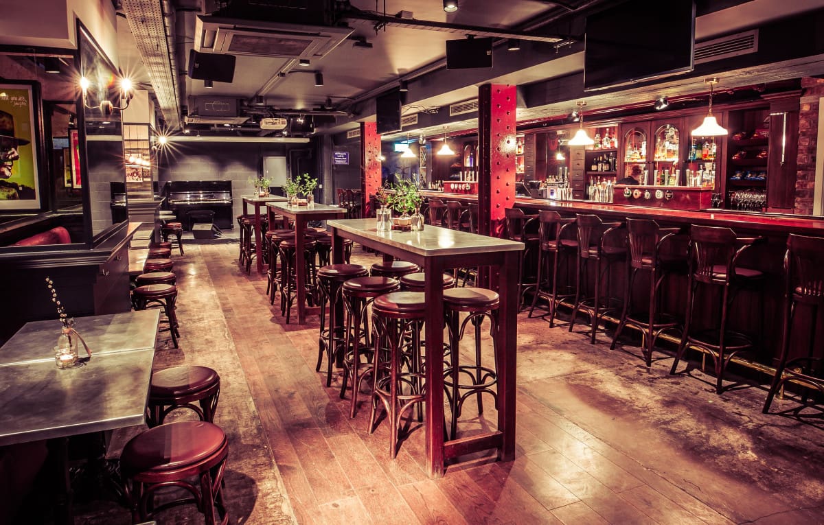 A warmly lit bar with high tables and stools, a long polished counter, hanging pendant lights, and shelves stocked with bottles. The wooden floor and red accents create a cosy, inviting atmosphere.