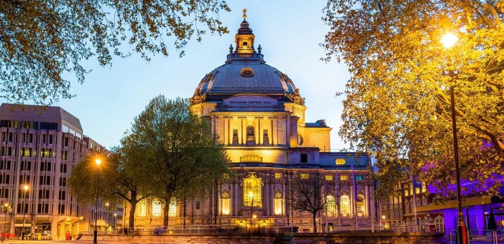 A grand historic building with a large dome and ornate architecture is illuminated at dusk, surrounded by trees and modern buildings, with streetlights glowing in the foreground.