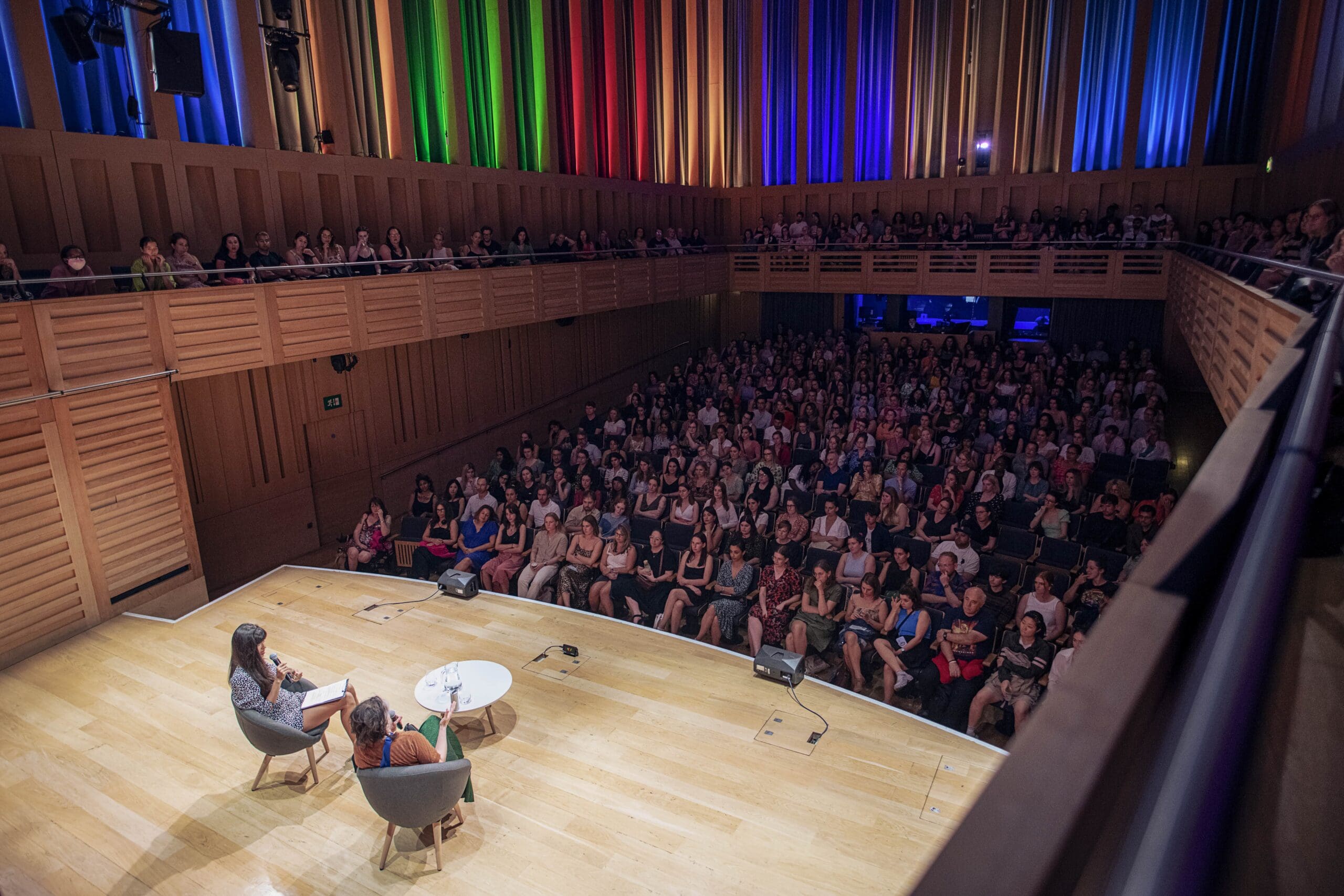 Two people sit on stage having a discussion in front of a large, seated audience in a modern auditorium. The walls have wooden panelling and vertical coloured lights in shades of blue, green, red, and yellow.