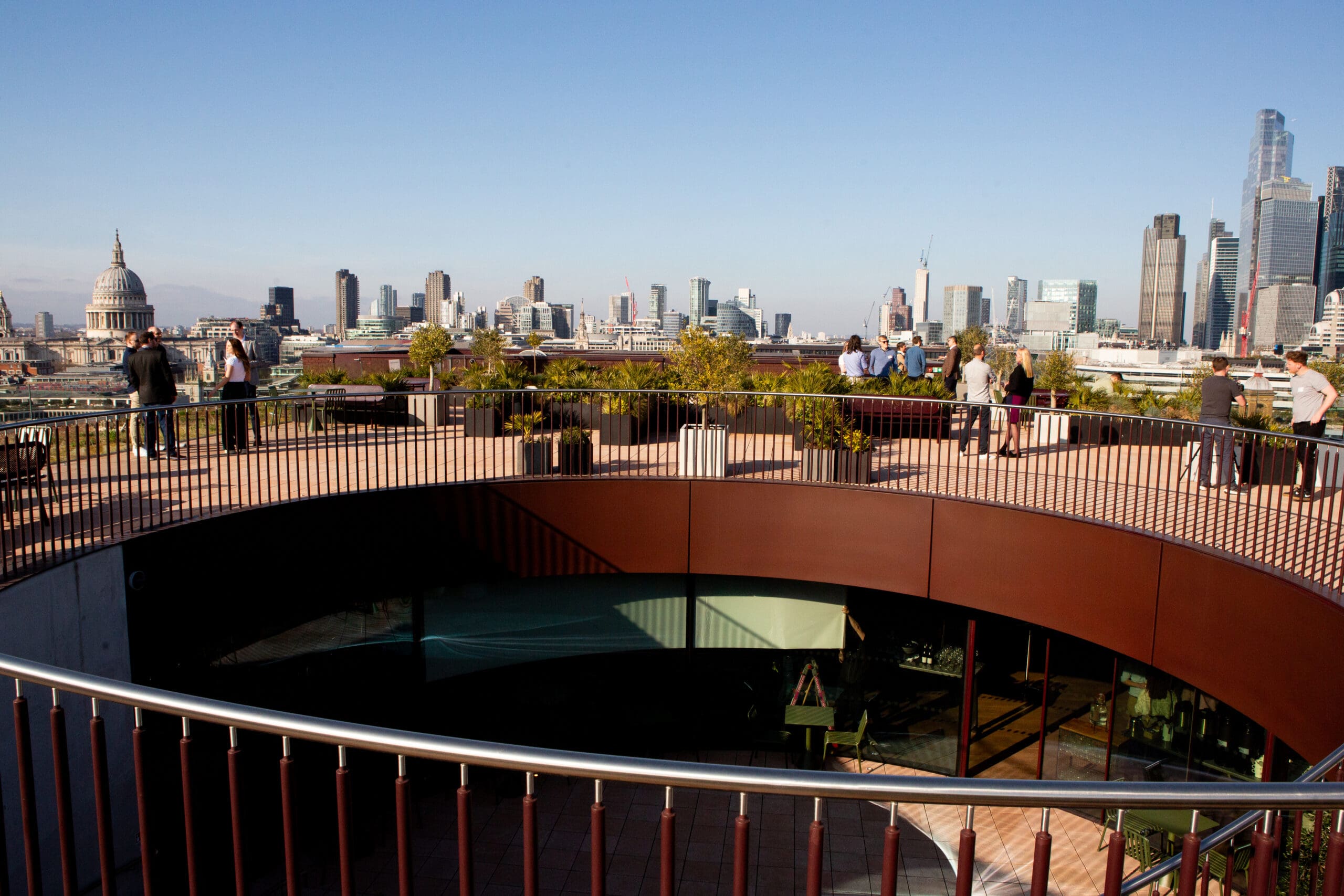 People stand on a circular rooftop terrace with plants, overlooking the London skyline, including St Paul’s Cathedral and modern skyscrapers under a clear sky.