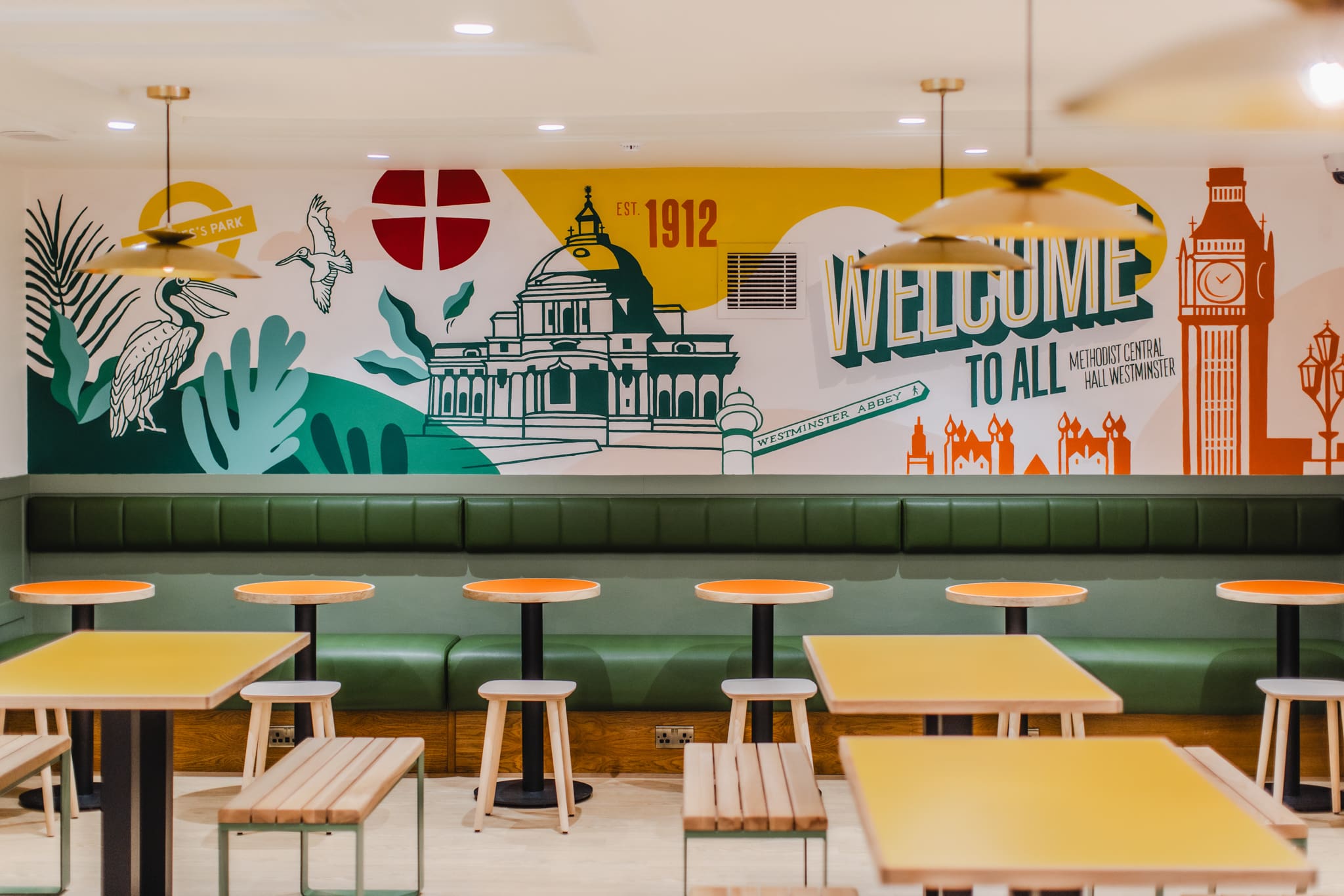 A modern dining area with yellow tables, wooden benches, green cushioned seating, and a colourful mural featuring city landmarks, birds, and bold text that says 