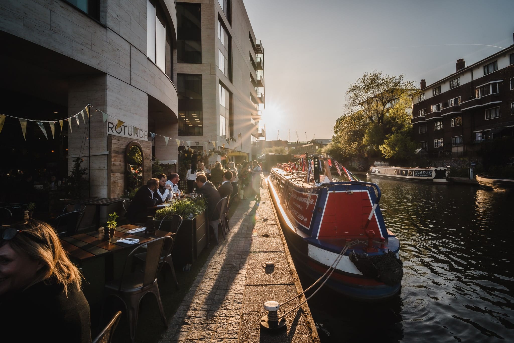 People dining outdoors beside a canal at sunset, with a red and blue canal boat moored nearby. Sunlight streams between modern and older buildings, creating a warm, lively atmosphere.