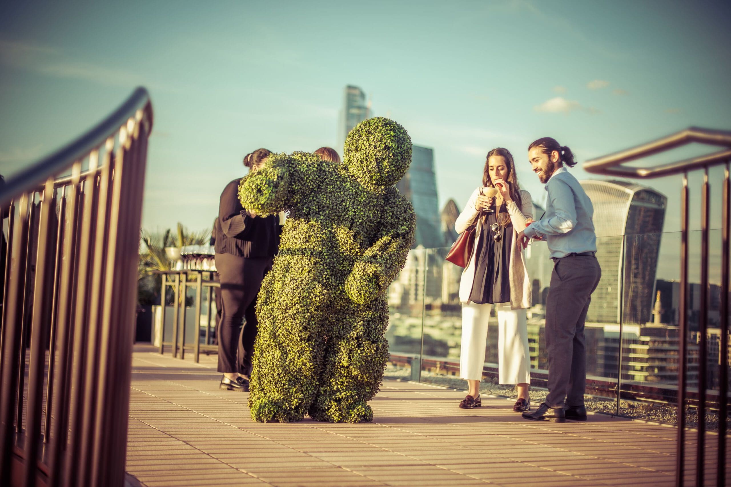 A person in a leafy green shrub costume entertains people on a rooftop terrace, while two individuals smile and look at a mobile. Modern city buildings are visible in the background under a clear sky.