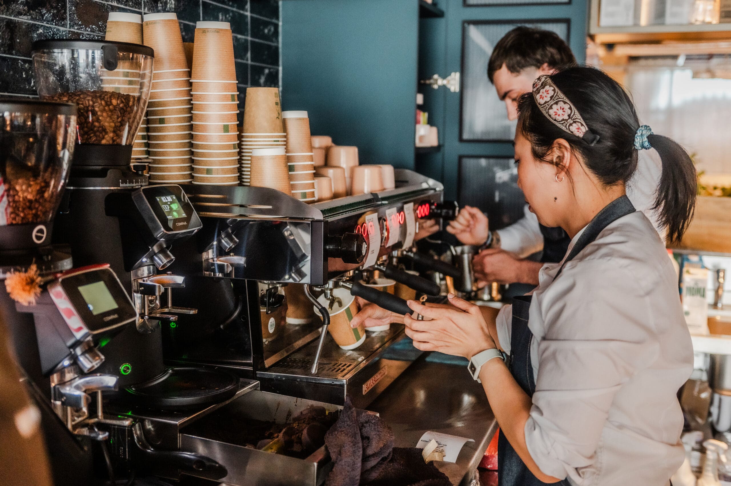Two baristas, one male and one female, prepare coffee drinks using an espresso machine in a busy café. Stacks of paper cups and coffee-making equipment are visible on the counter.