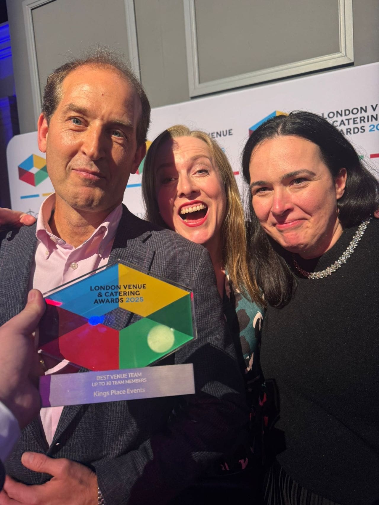 Three smiling people pose together at the London Venue & Catering Awards 2023, holding a colourful trophy for Kings Place Events. Event branding is visible on the backdrop behind them.