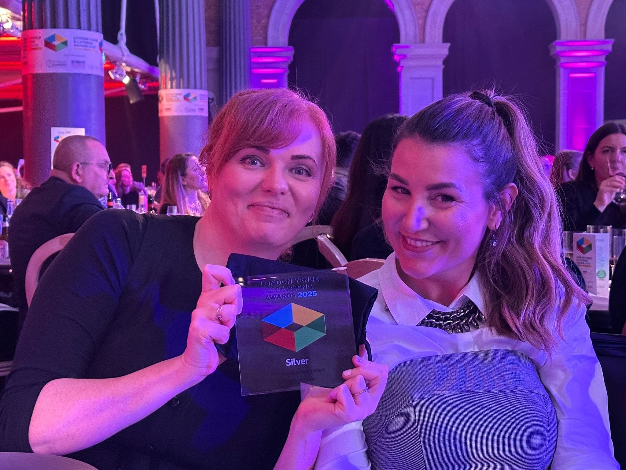 Two smiling women sit at a table holding a glass trophy that reads “Silver.” They are dressed formally, and the background shows other people seated at tables in a venue lit with purple lights.