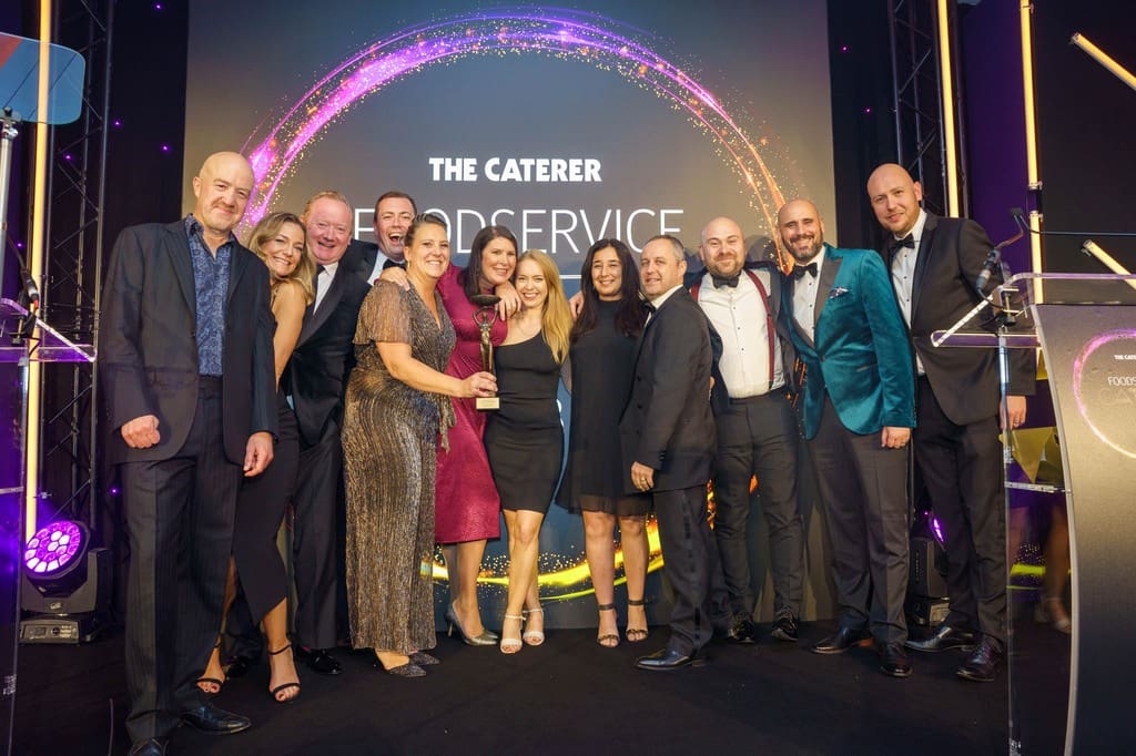 A group of people dressed in formal attire stand together on stage, smiling and posing for a photo at The Caterer Foodservice awards event, with colourful lights and a large backdrop behind them.