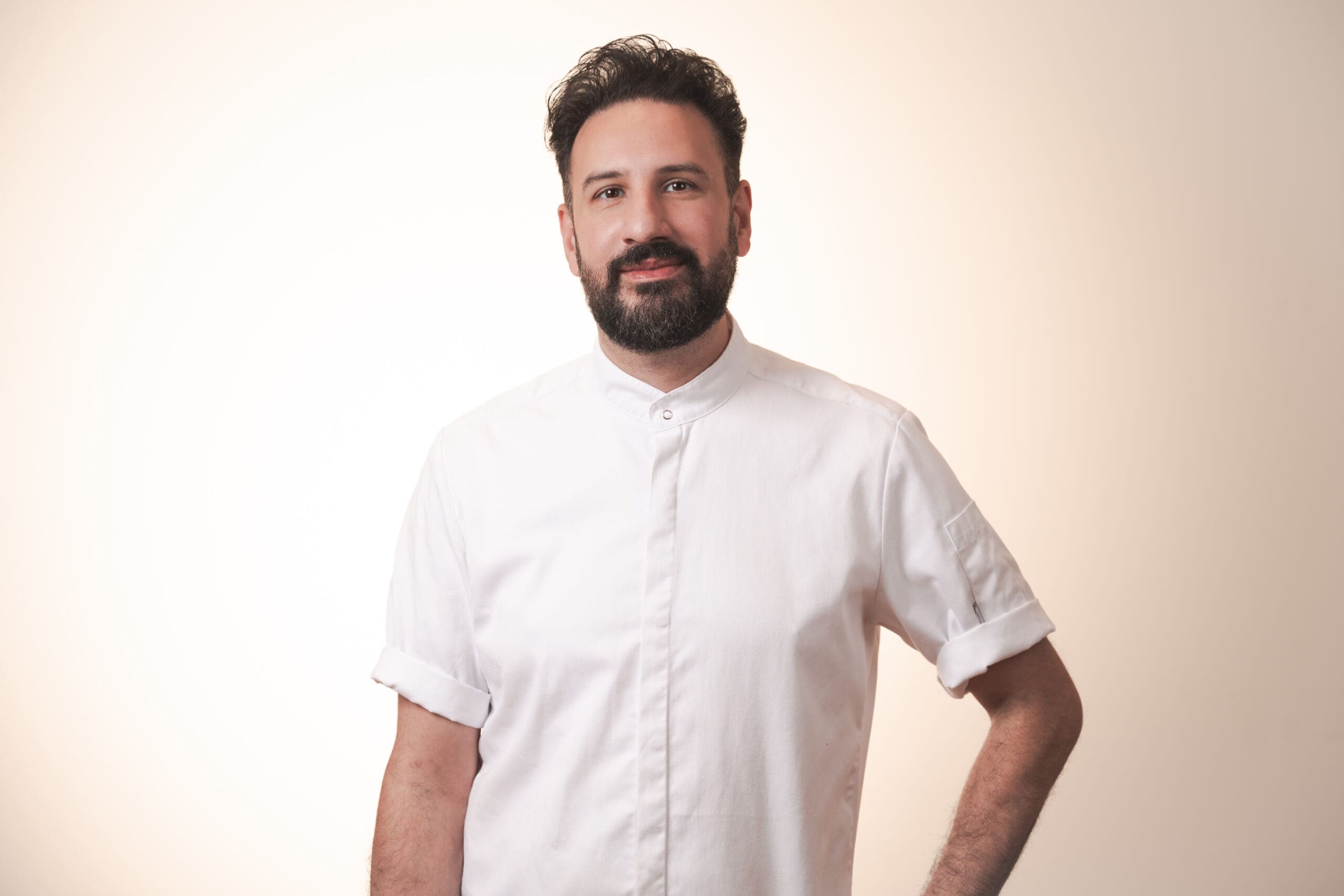 A man with dark hair and a beard is standing against a light background, wearing a white buttoned shirt with rolled-up sleeves. He is smiling softly and has one hand on his hip.