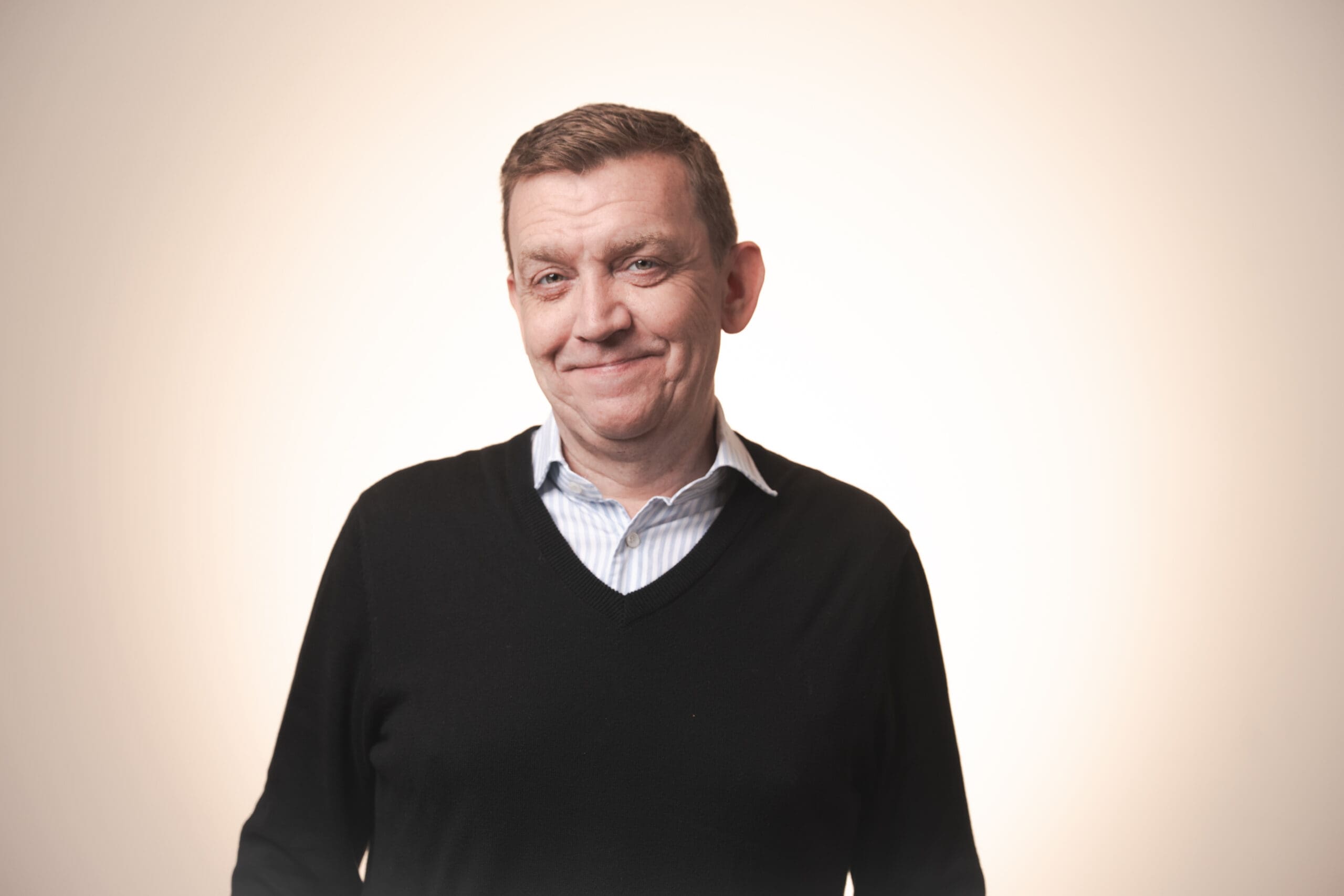 A middle-aged man with short brown hair, wearing a black jumper over a collared shirt, stands against a light, neutral background, smiling slightly at the camera.