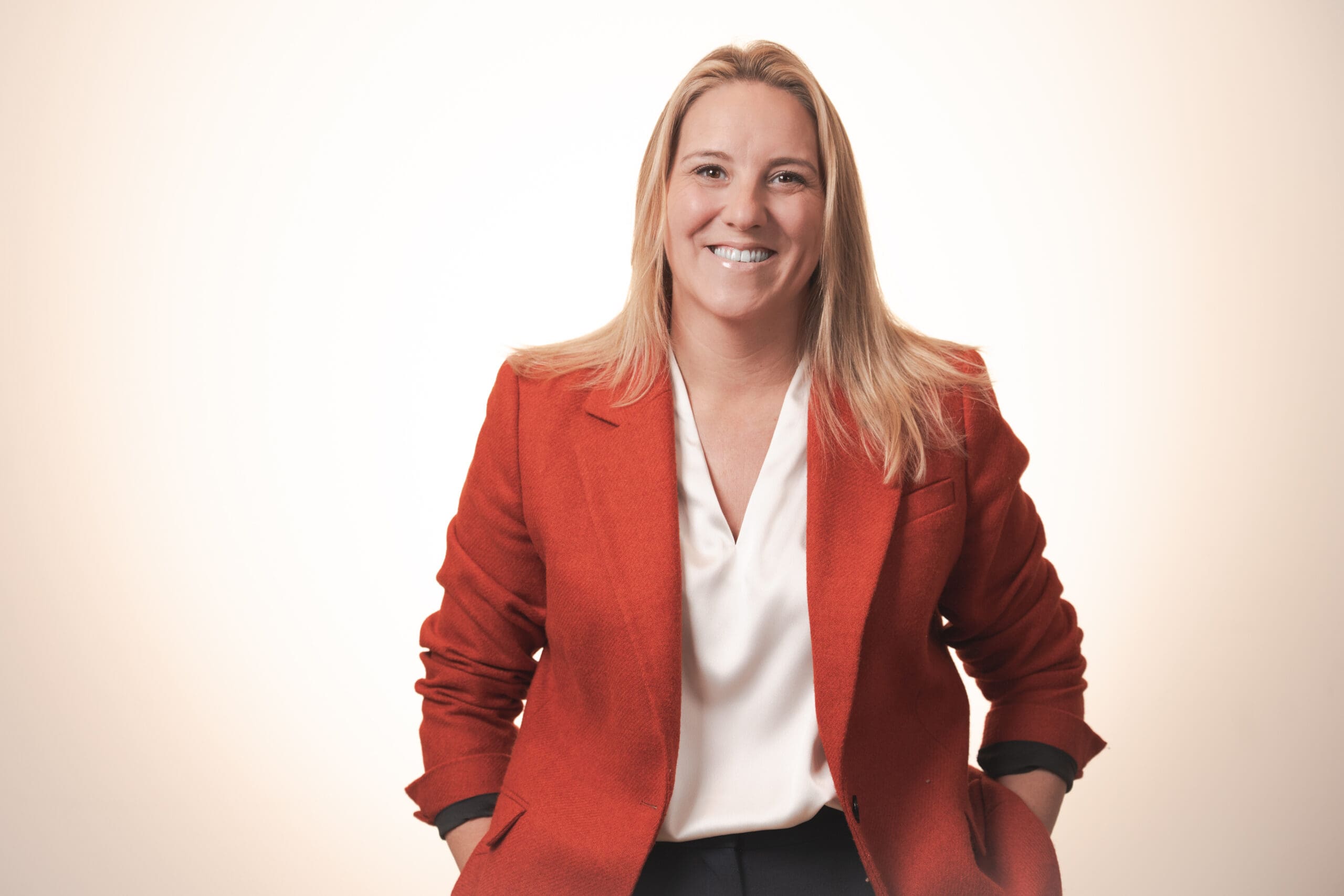 A smiling woman with straight blonde hair wears a bright red blazer over a white blouse, sitting with her hands resting on her legs against a light, neutral background.