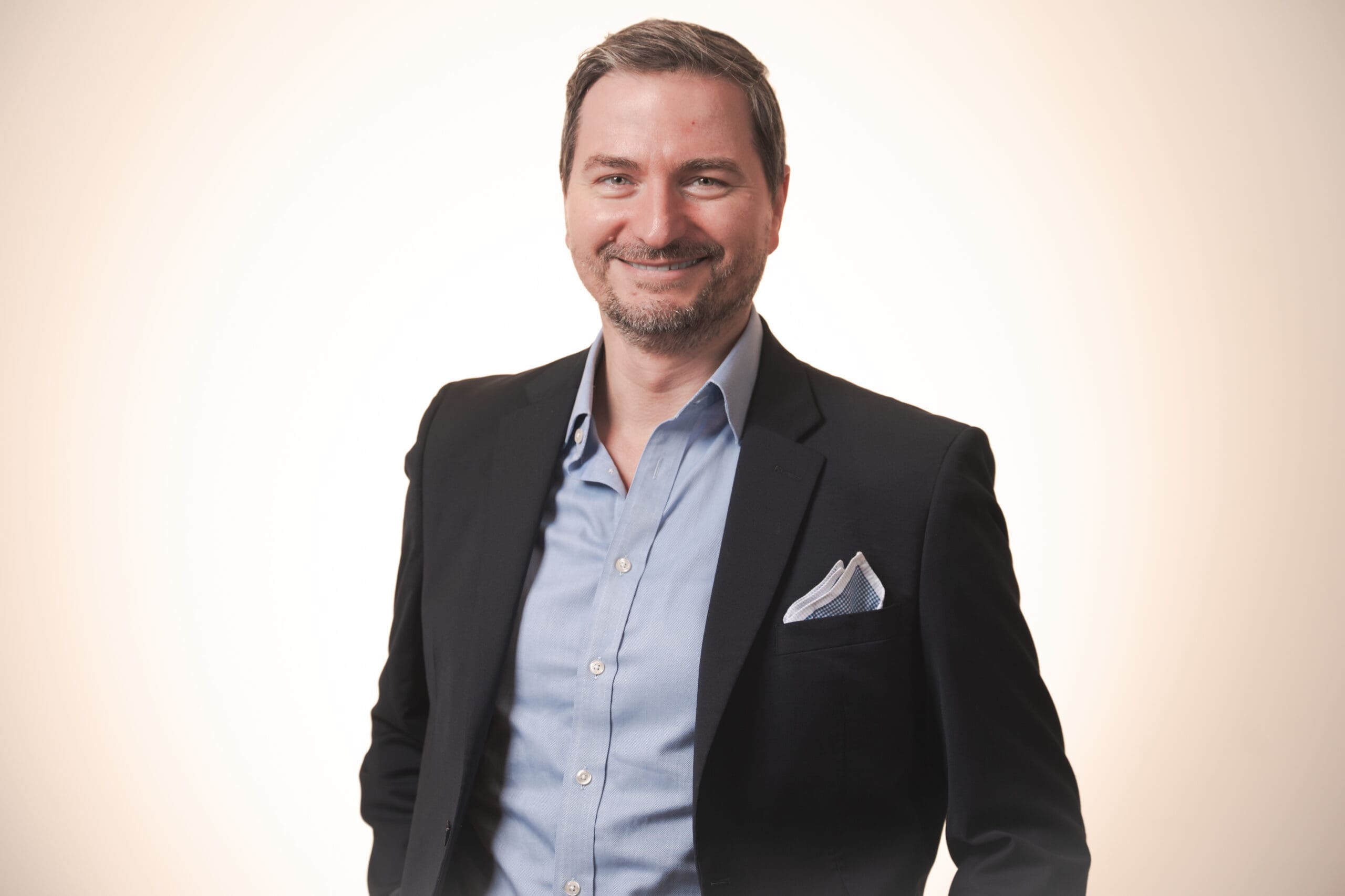 A man in a dark blazer with a light blue dress shirt and pocket square smiles confidently against a plain light background.