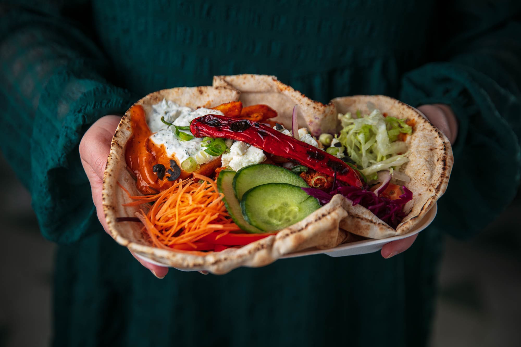 A person in a green dress holds a plate with pitta bread filled with colourful vegetables, including sliced cucumber, shredded carrots, lettuce, red cabbage, grilled pepper, and a dollop of creamy sauce.