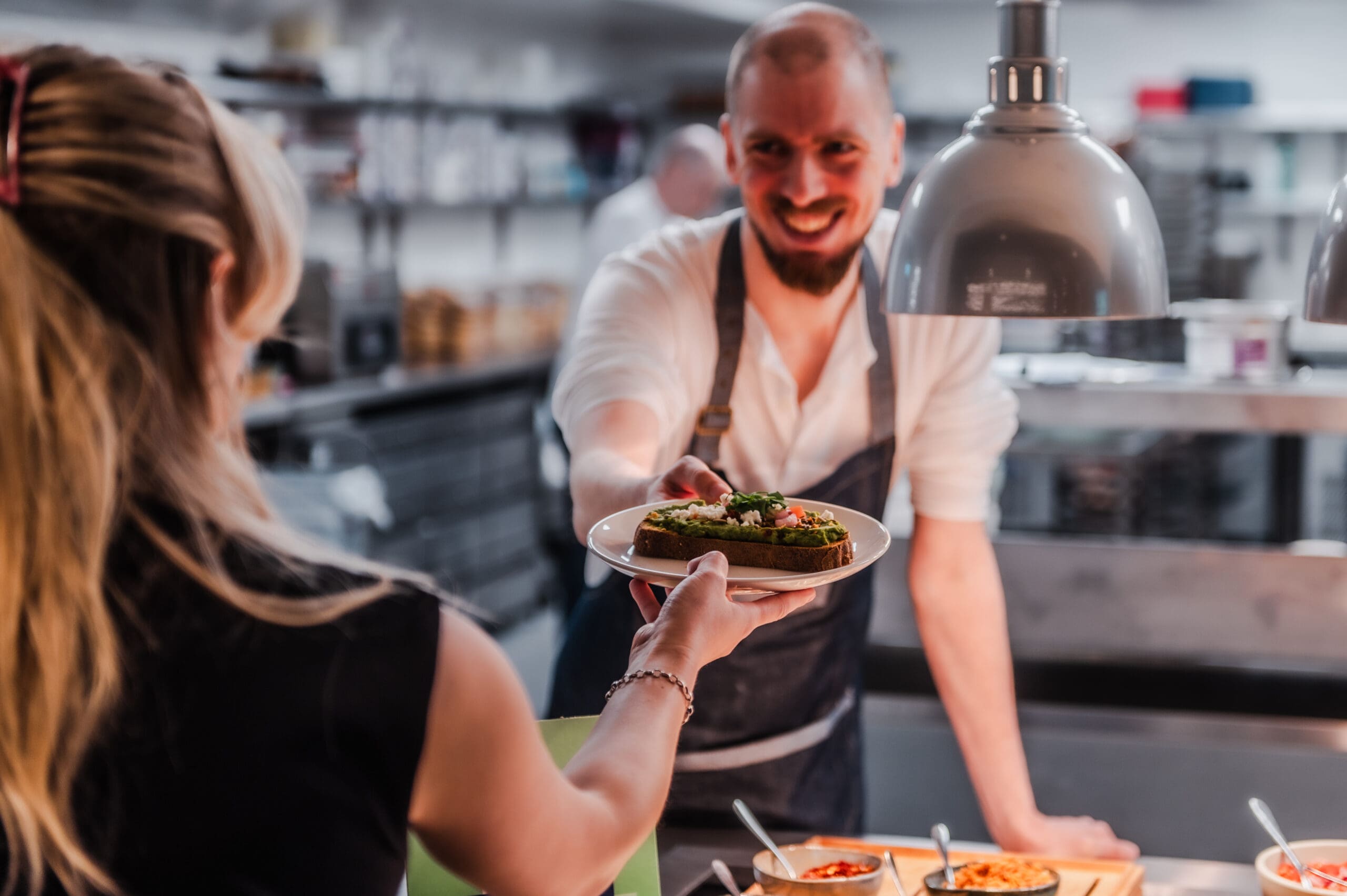 A smiling chef in an apron hands a plated dish to a customer across the kitchen counter, with various food items visible in serving trays nearby.