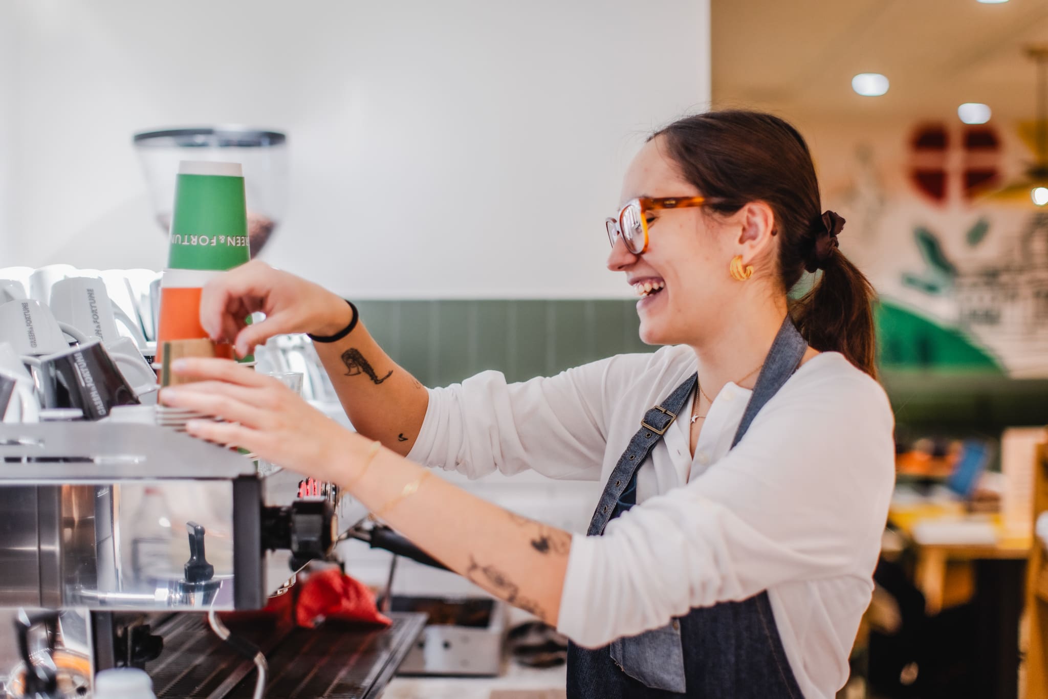 A smiling barista with glasses and tattoos prepares a coffee drink behind a café counter, placing a green and orange cup on an espresso machine.