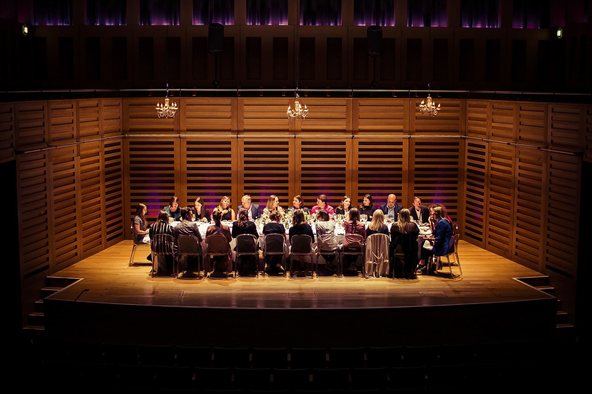 A large group of people sit around a long table on a stage in a wooden-panelled concert hall, lit by warm overhead lights and chandeliers, with empty audience seats in the foreground.