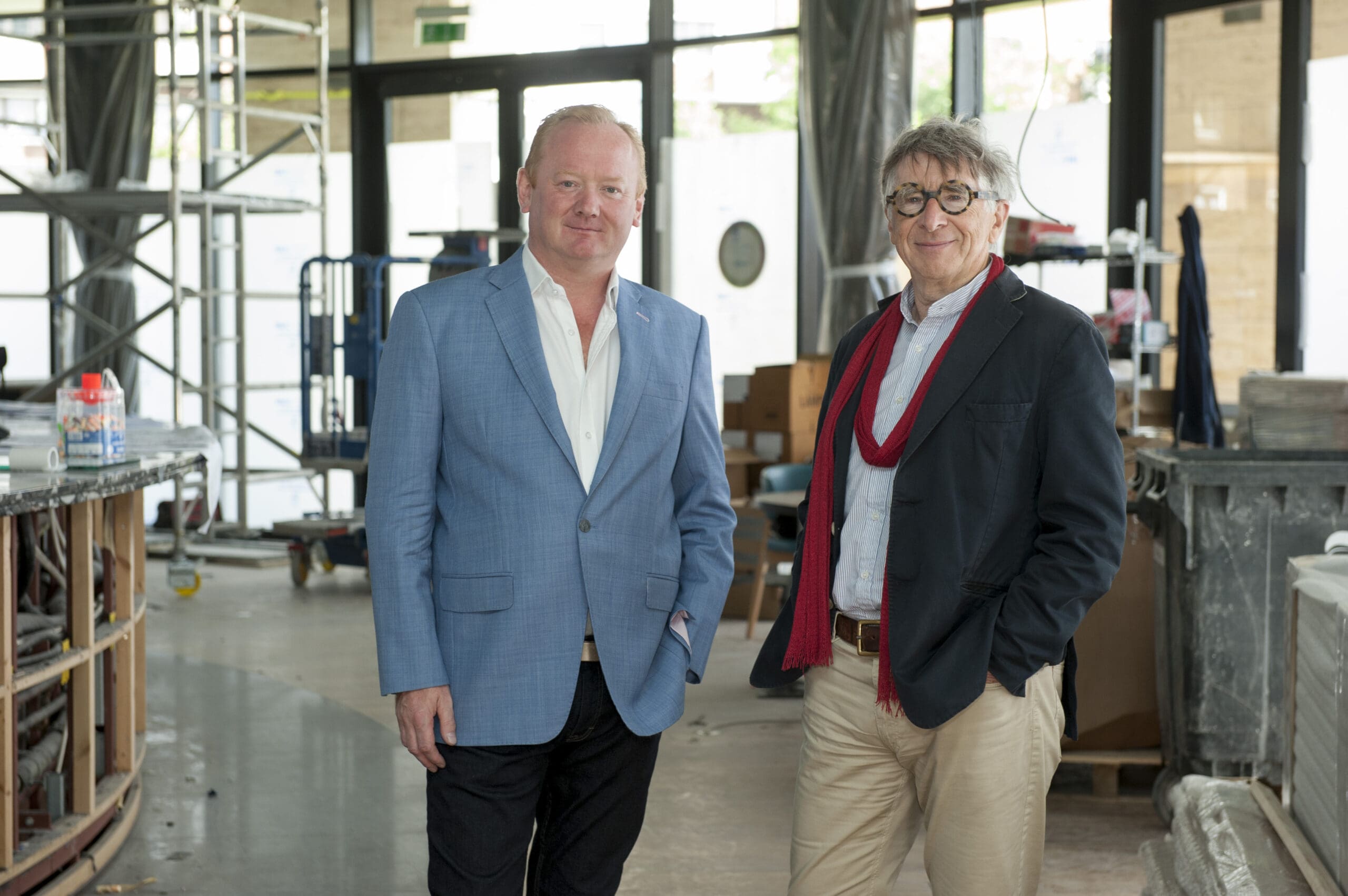 Two men stand side by side inside a spacious, unfinished building. One wears a light blue blazer, the other a dark jacket with a red scarf. Construction materials and equipment are visible in the background.