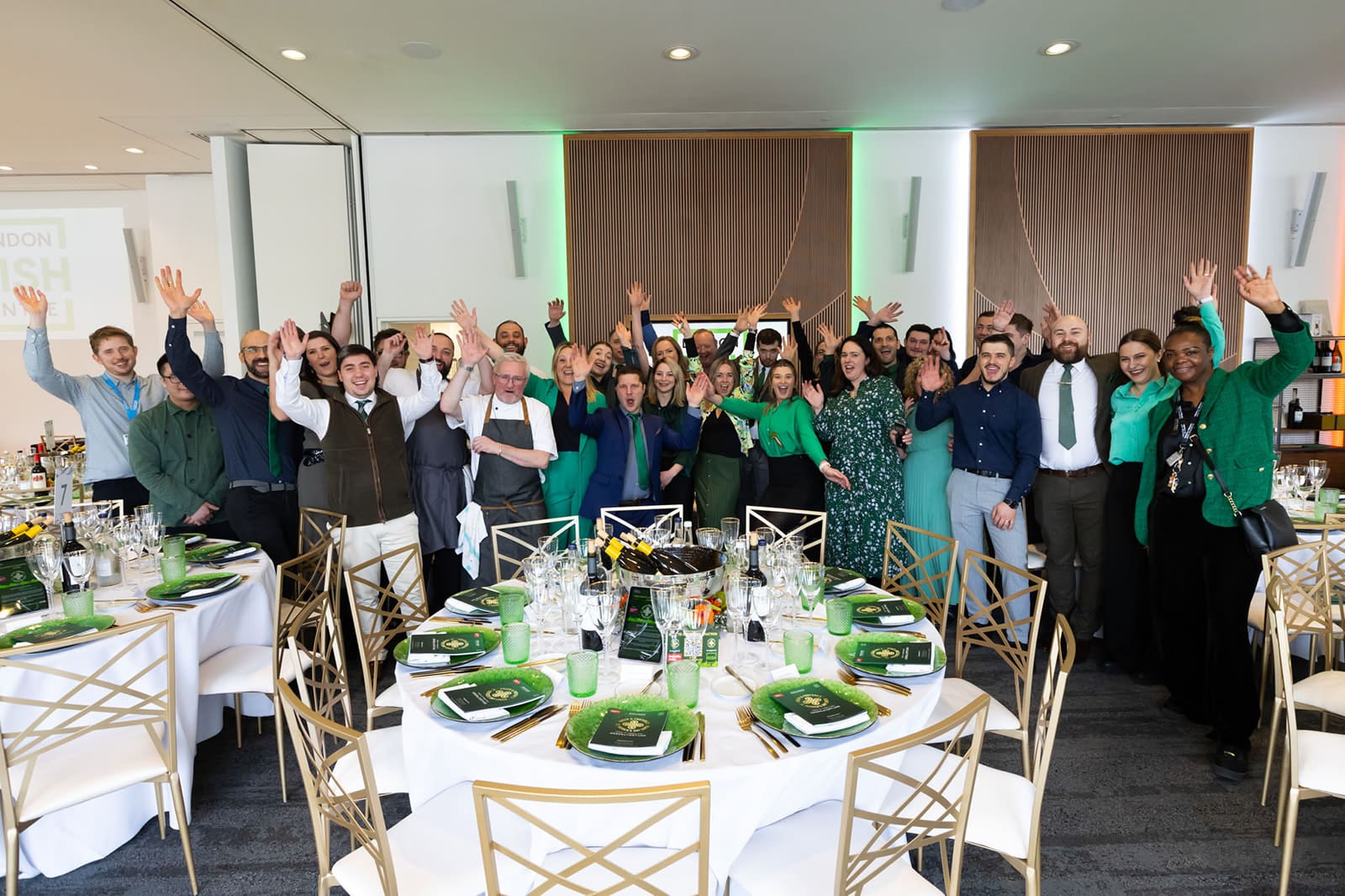 A large group of people dressed in green and formal attire stand together, smiling and raising their hands in celebration around elegantly set tables at an indoor event.