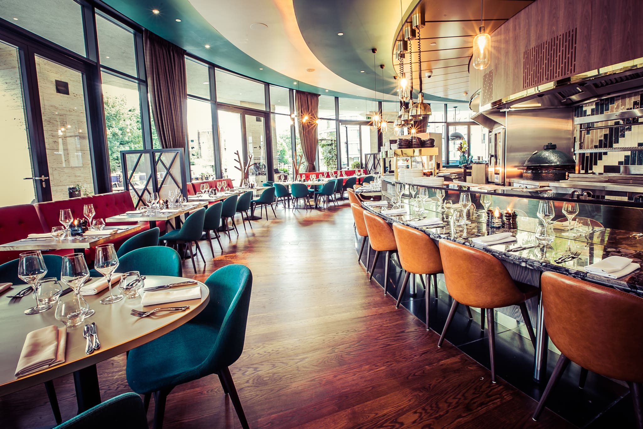 Modern restaurant interior with elegant tables set for dining, upholstered teal and orange chairs, large windows letting in natural light, and an open kitchen area with contemporary decor and wooden floors.