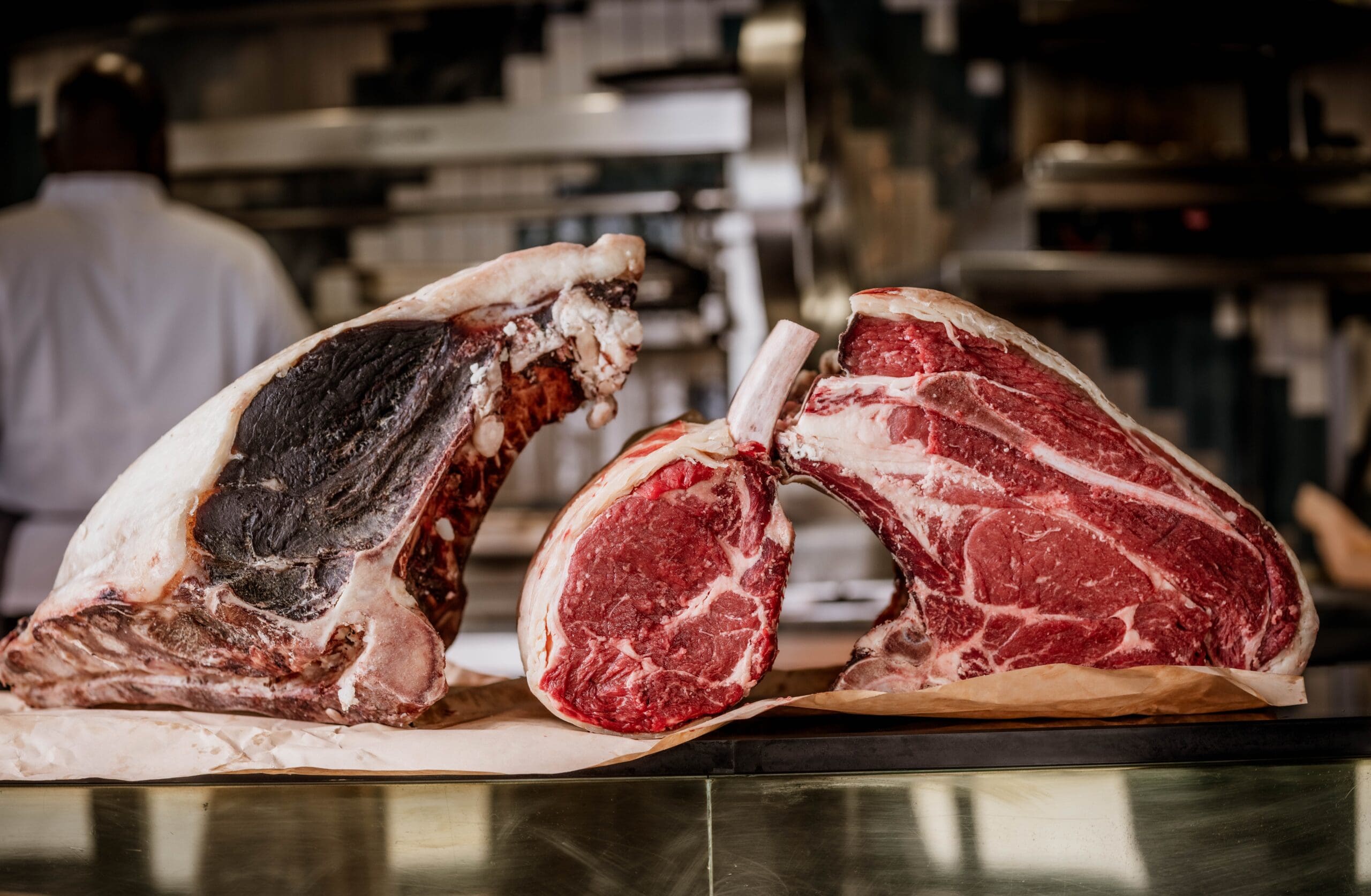 Two large raw cuts of beef, including a dry-aged steak with darkened outer edges, sit on butcher’s paper atop a metal counter in a professional kitchen. A chef in a white coat is blurred in the background.