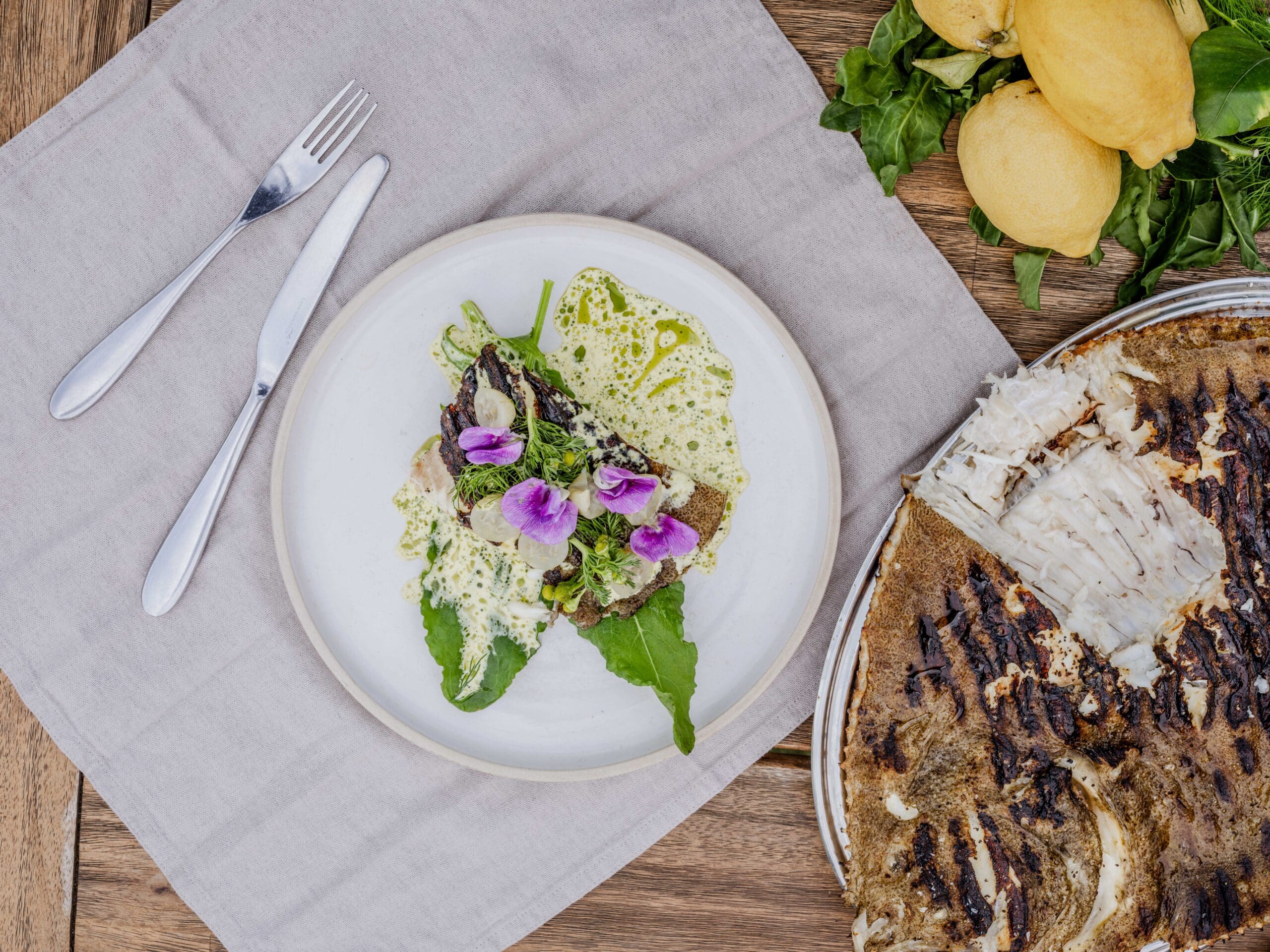 A plated gourmet dish with green sauce, leafy greens, and edible flowers sits on a linen serviette next to a set of fork and knife; nearby are lemons, greens, and a partially cut grilled fish on a wooden table.