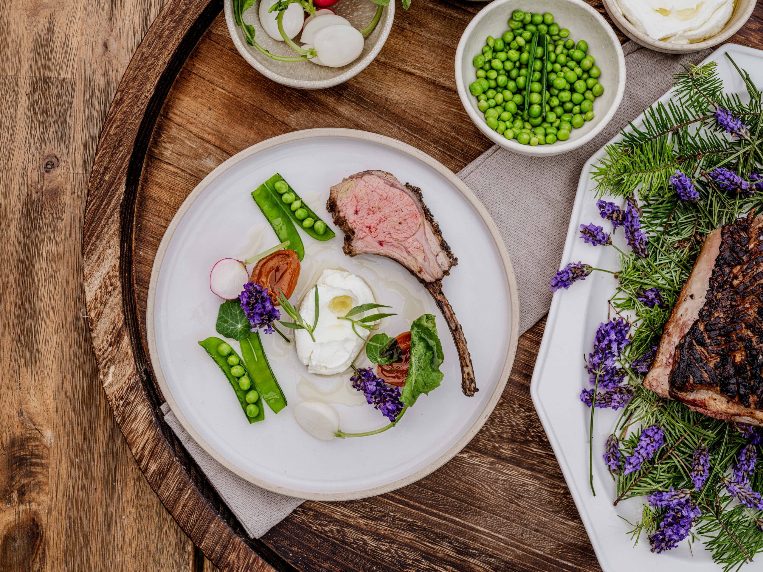 A plated lamb chop with fresh peas, radish slices, greens, and edible purple flowers, served with a creamy white sauce. Surrounding bowls contain peas and other fresh vegetables on a rustic wooden tray.