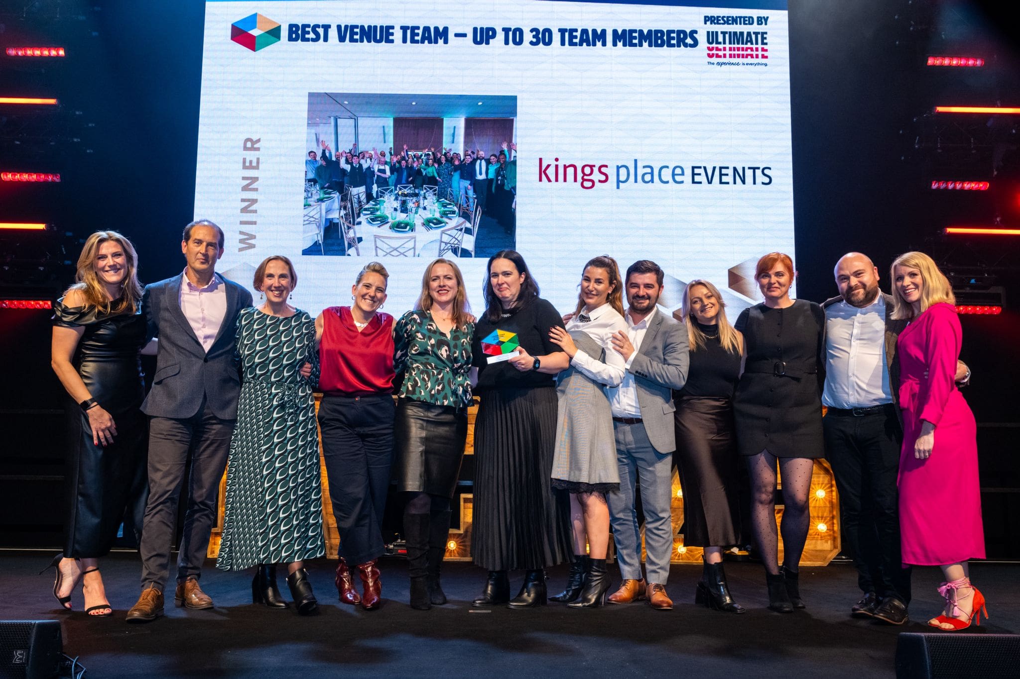 A group of people stand smiling on stage, holding an award. Behind them, a screen displays Best Venue Team – Up to 30 Team Members and kings place EVENTS alongside a photo of a banquet.