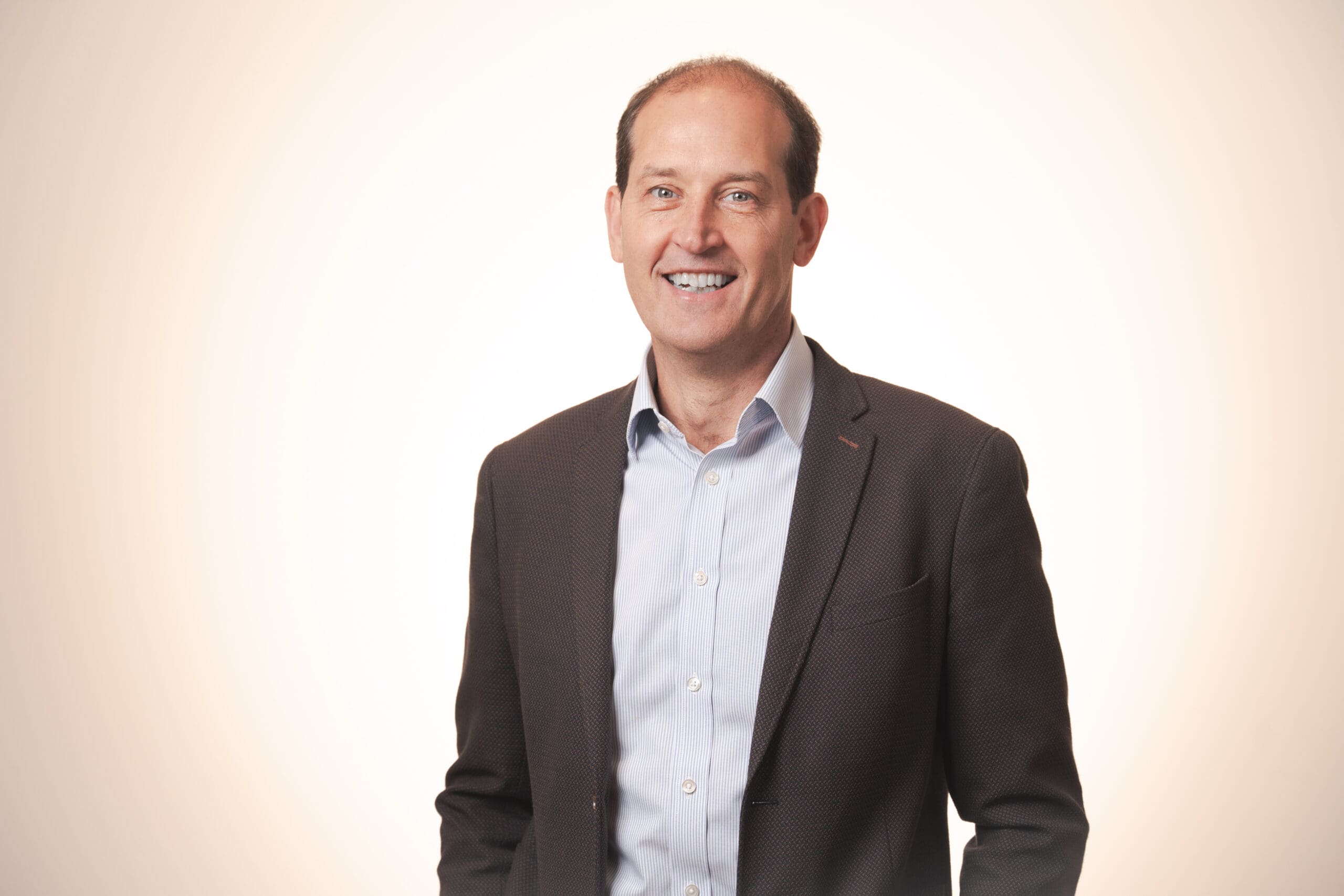 A man in a business suit is smiling and standing against a plain light background. He is wearing a dark jacket over a light blue shirt and has short brown hair.