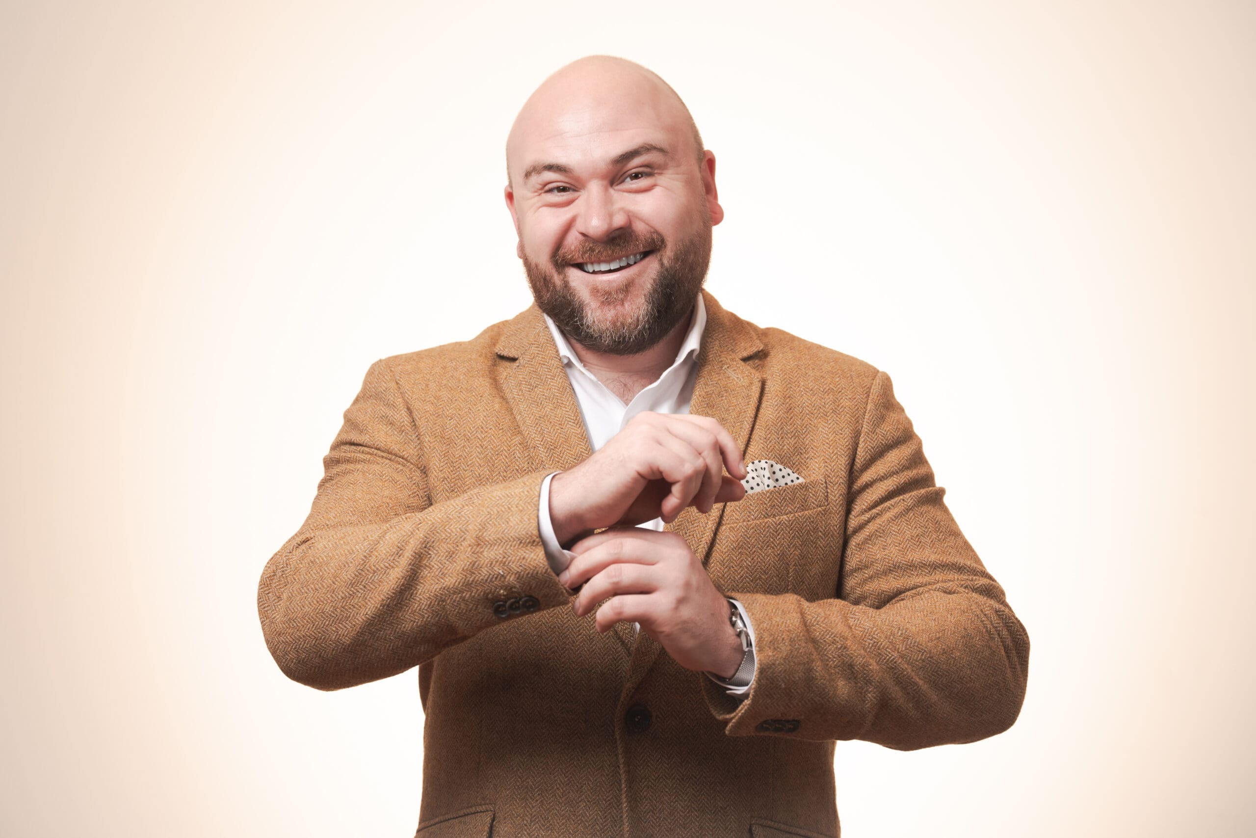A smiling bald man with a beard wearing a brown blazer and white shirt adjusts his pocket handkerchief against a soft beige background.