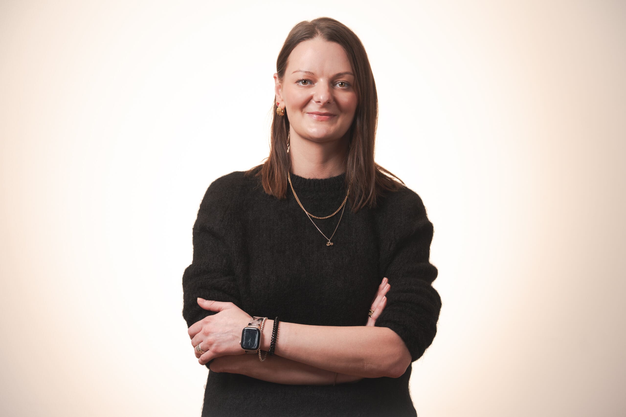 A woman with straight brown hair and light skin stands smiling with arms folded. She wears a black jumper, layered necklaces, bracelets, and a smartwatch, set against a light, neutral background.