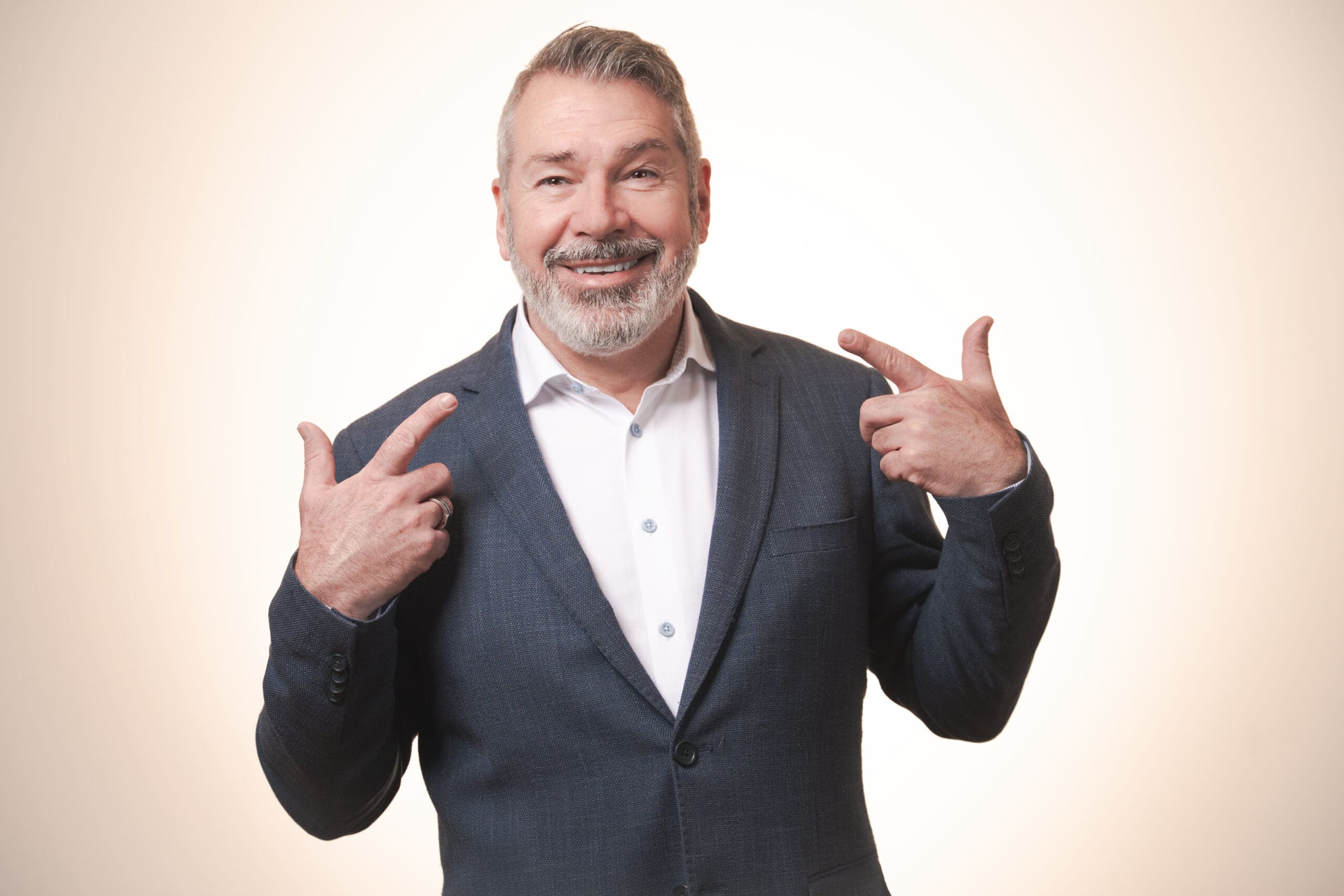 A middle-aged man with grey hair and a beard, wearing a dark blazer and white shirt, smiles and points at himself with both hands against a light background.