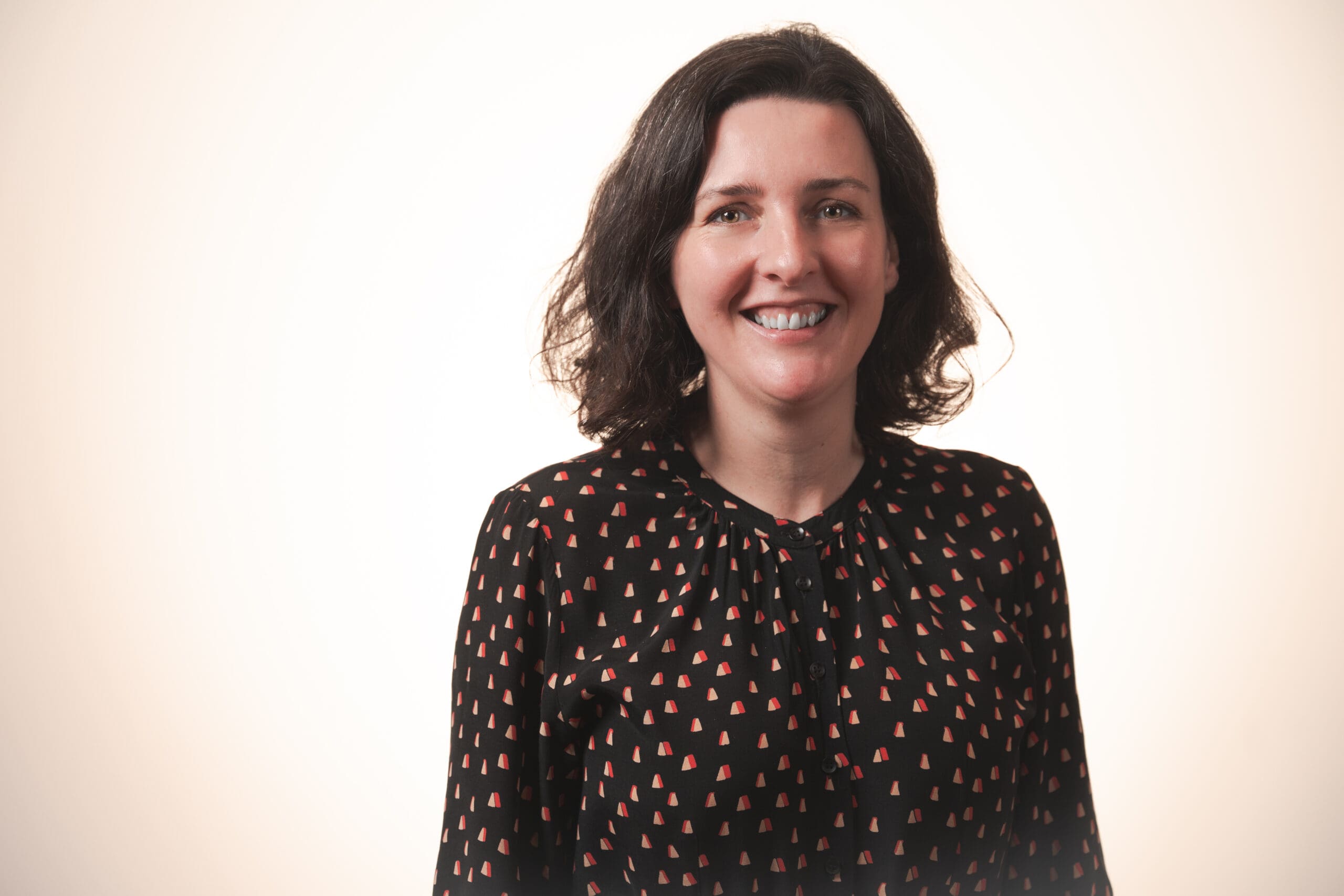 A woman with shoulder-length dark hair smiles at the camera. She is wearing a black blouse with a pattern of red and white triangles, standing against a plain, light-coloured background.