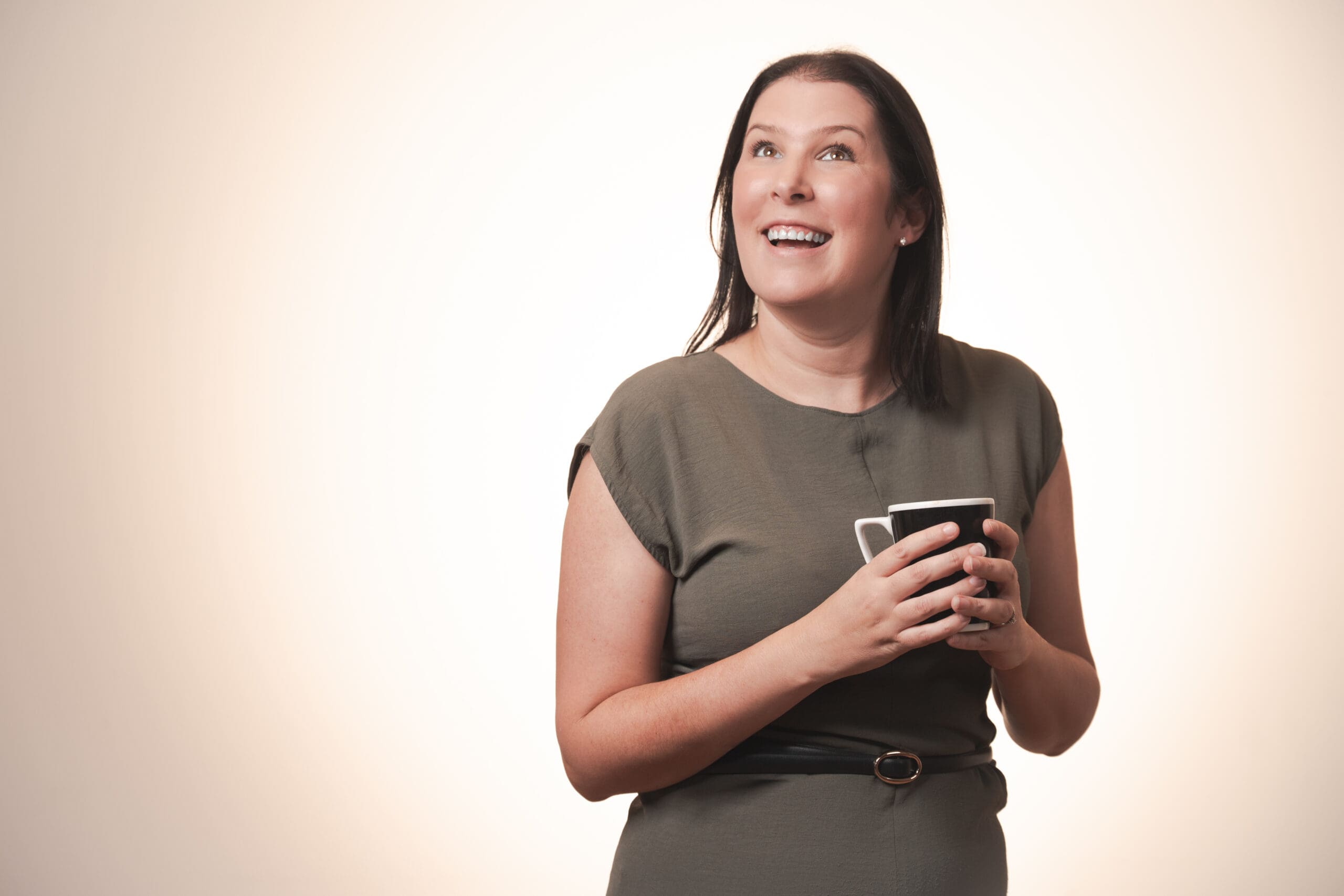 A woman with long dark hair, wearing a short-sleeved olive green dress, smiles and looks upwards while holding a black mug with both hands against a plain light background.