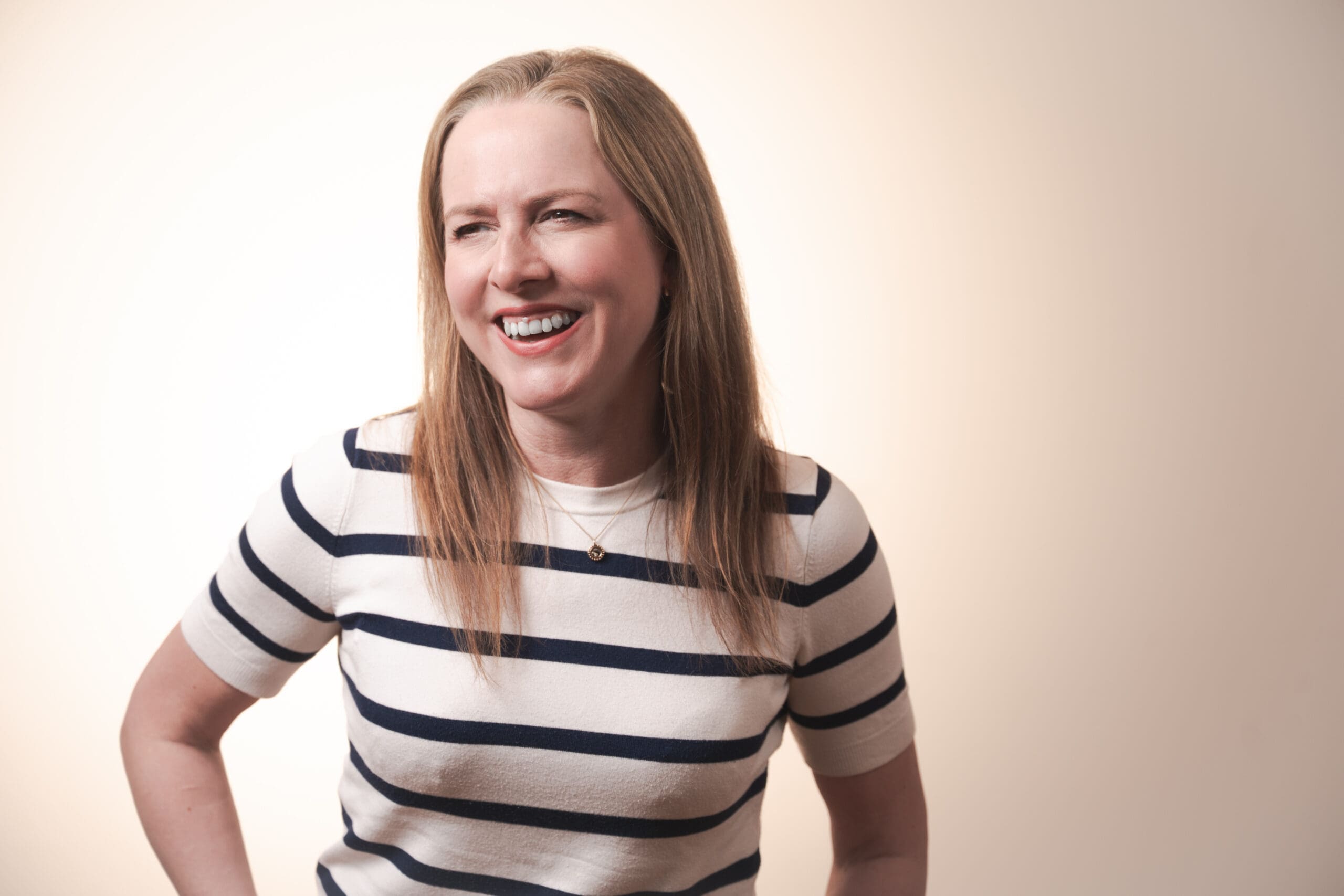 A woman with long blonde hair wearing a white shirt with black horizontal stripes smiles and laughs whilst looking slightly to the side against a neutral background.
