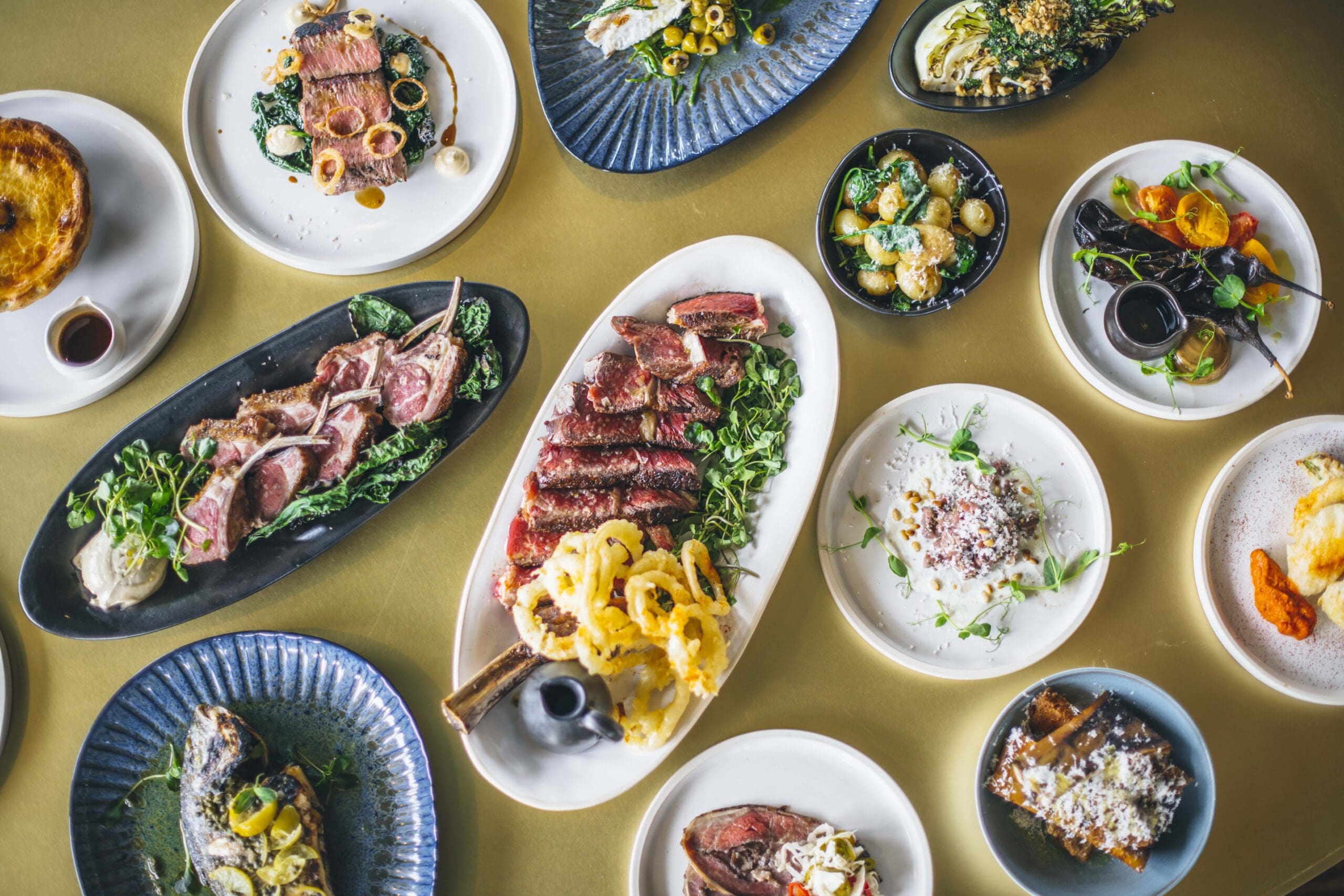 An assortment of gourmet dishes displayed on a table, including grilled meats, fish, vegetables, salads, and side dishes, all beautifully plated on variously shaped plates and bowls.