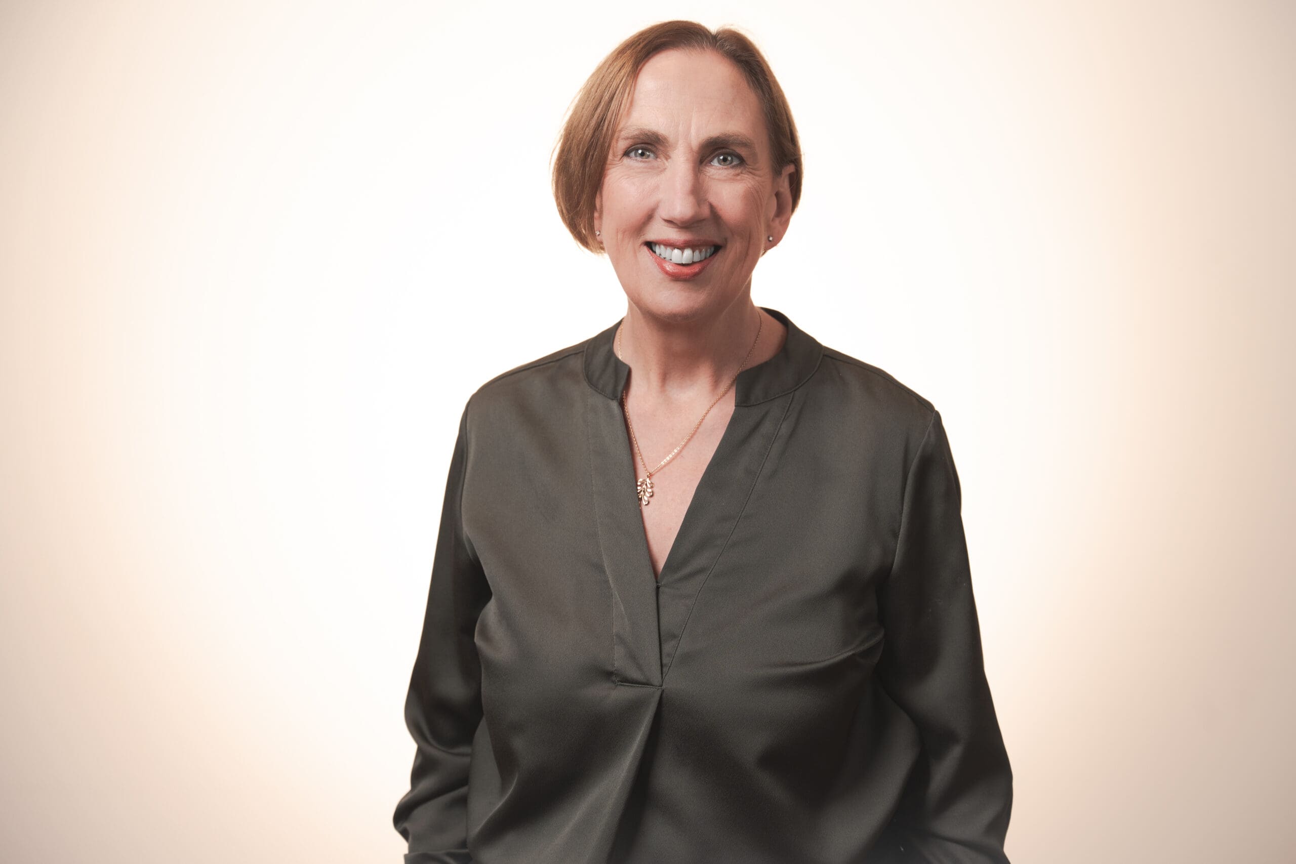 A smiling woman with short light brown hair wears a dark green blouse and a gold necklace, standing against a softly lit neutral background.