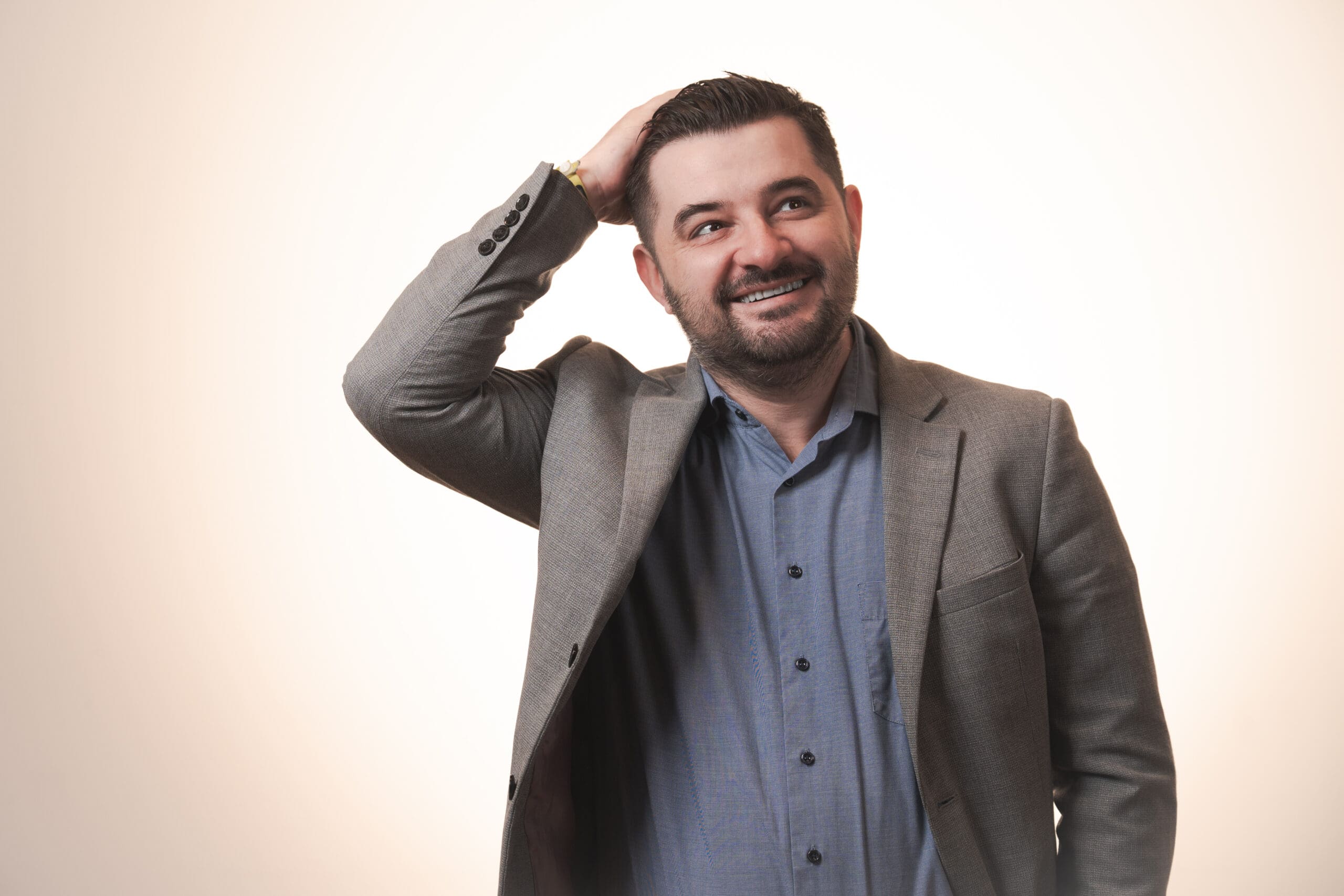 A man in a grey suit jacket and blue shirt stands against a light background, smiling and looking slightly upward while scratching his head with his right hand.