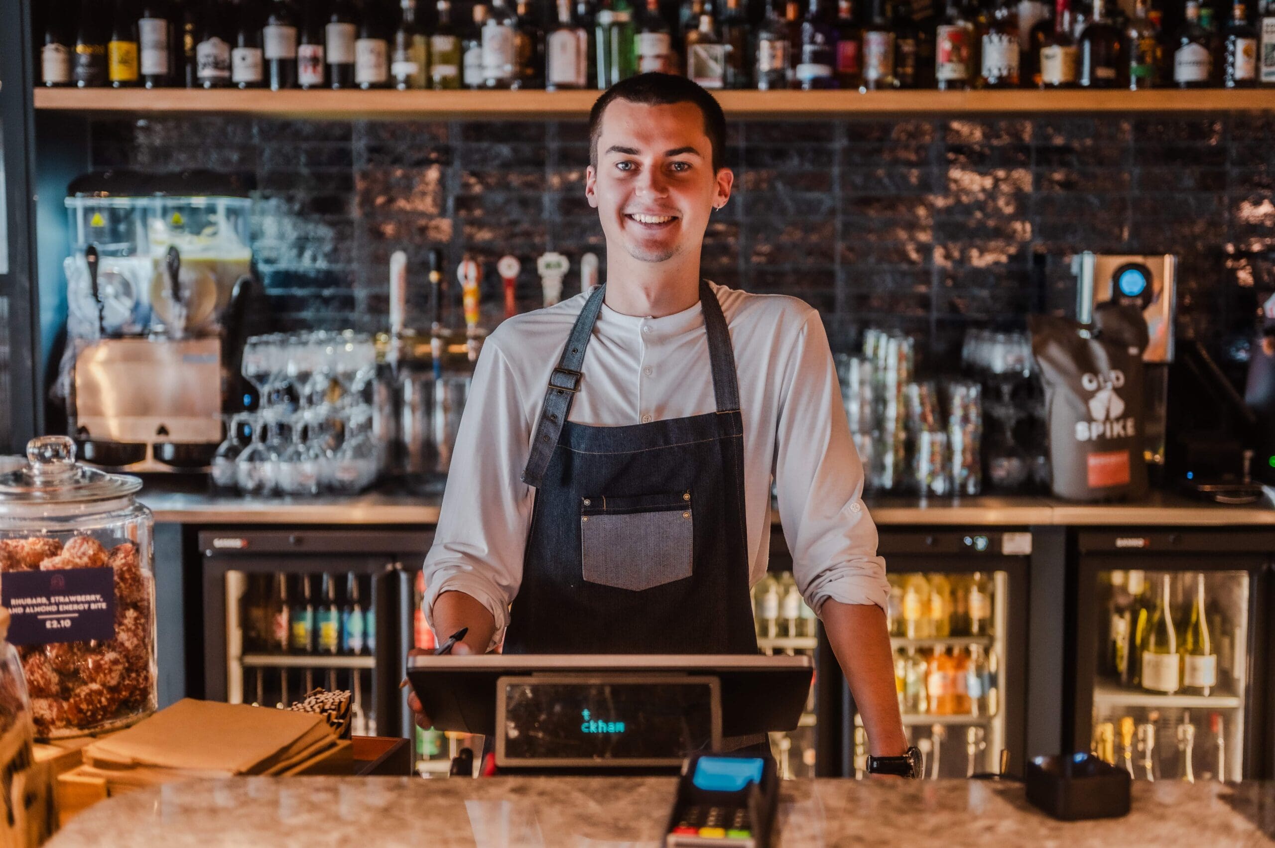A smiling barista wearing a dark apron stands behind the counter at a modern café, with shelves of bottles, glasses, and a coffee machine in the background. A till and various items are on the counter.