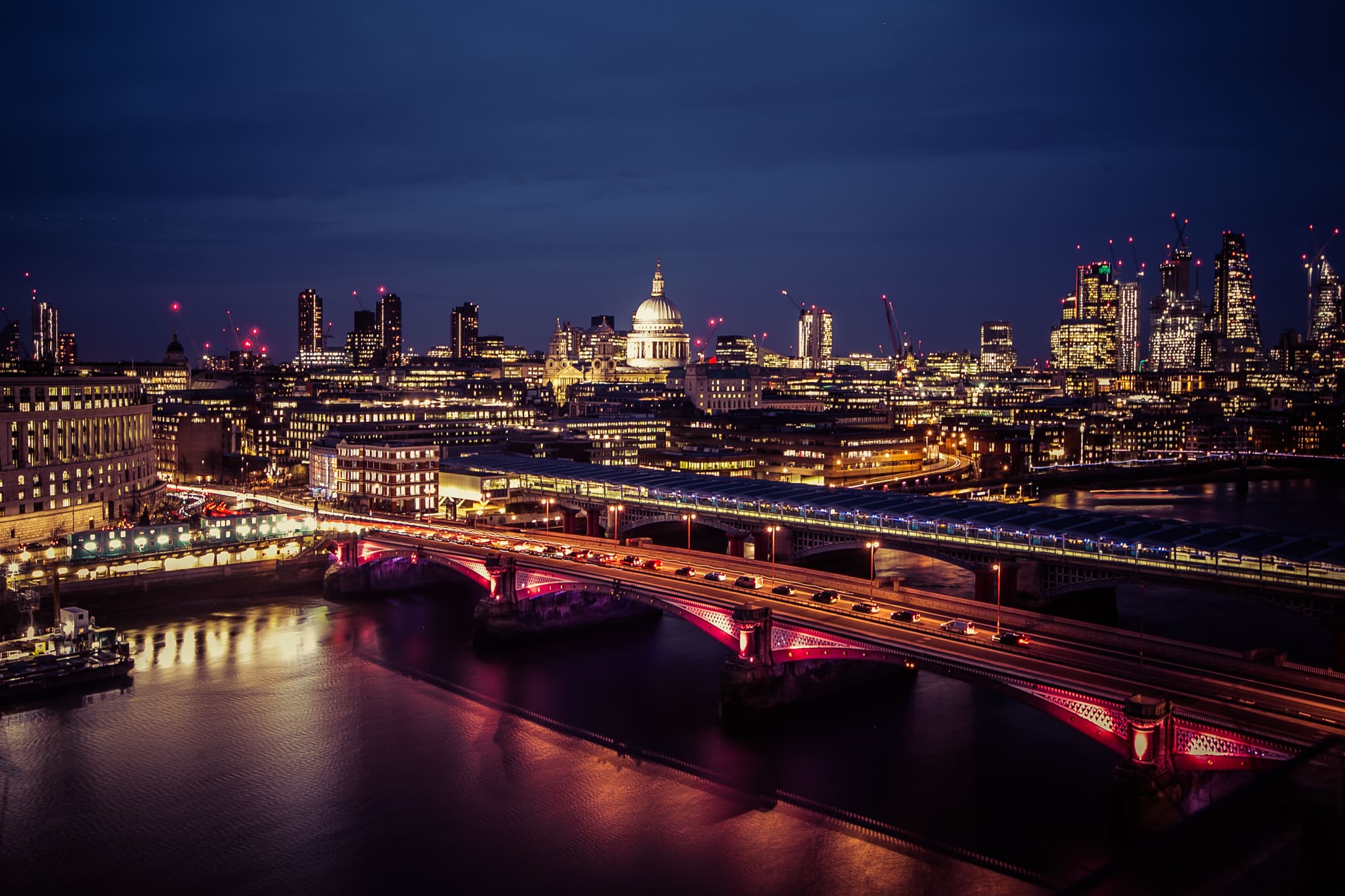 Night view of London with illuminated buildings, St Paul's Cathedral in the centre, and bridges over the River Thames. City lights reflect on the water, and the skyline includes modern skyscrapers and construction cranes.