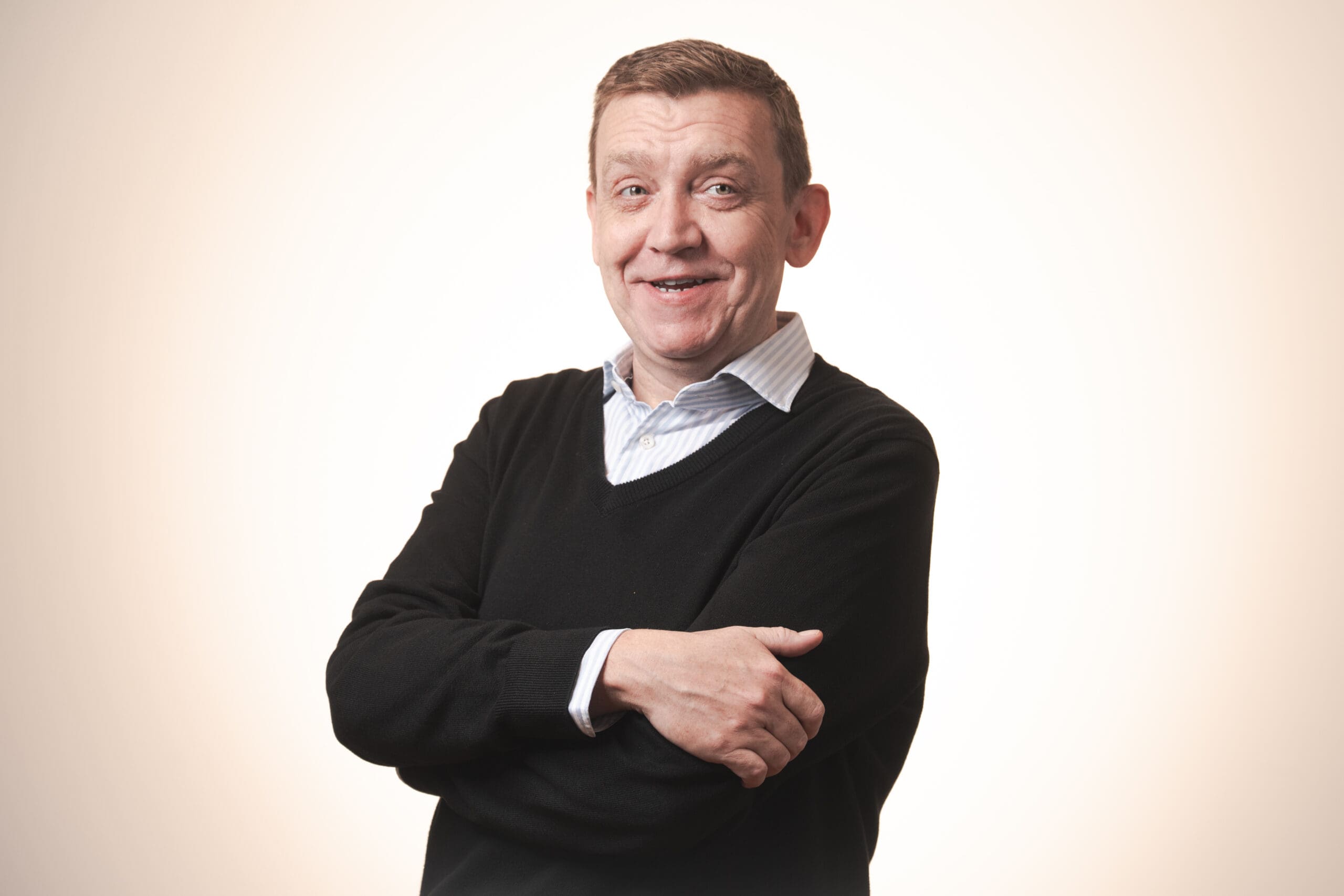 A middle-aged man with short brown hair smiles at the camera, standing with arms folded. He wears a black jumper over a collared shirt against a plain, light background.