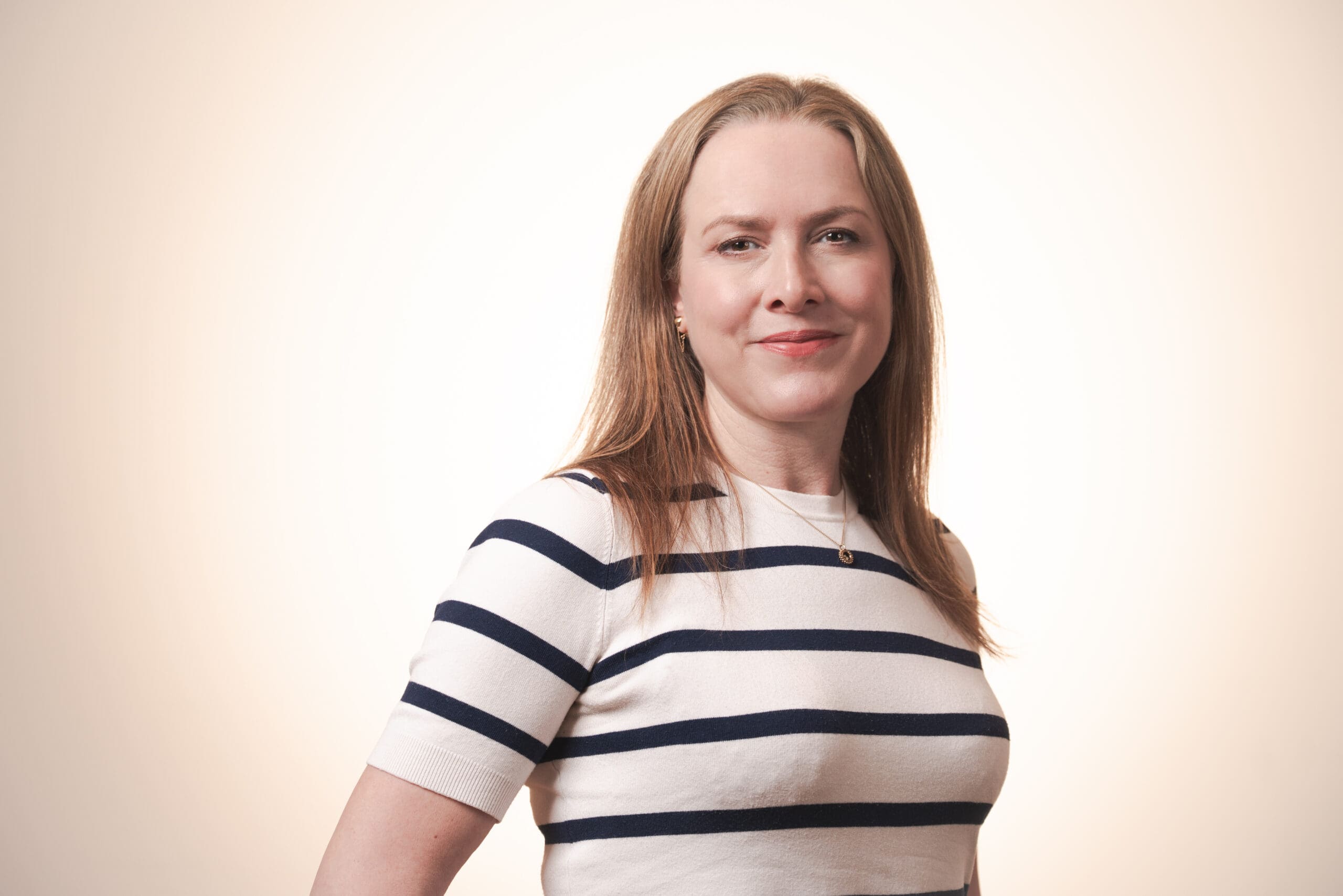 A woman with straight, light brown hair wearing a white shirt with navy horizontal stripes stands against a softly lit, neutral background, smiling slightly at the camera.