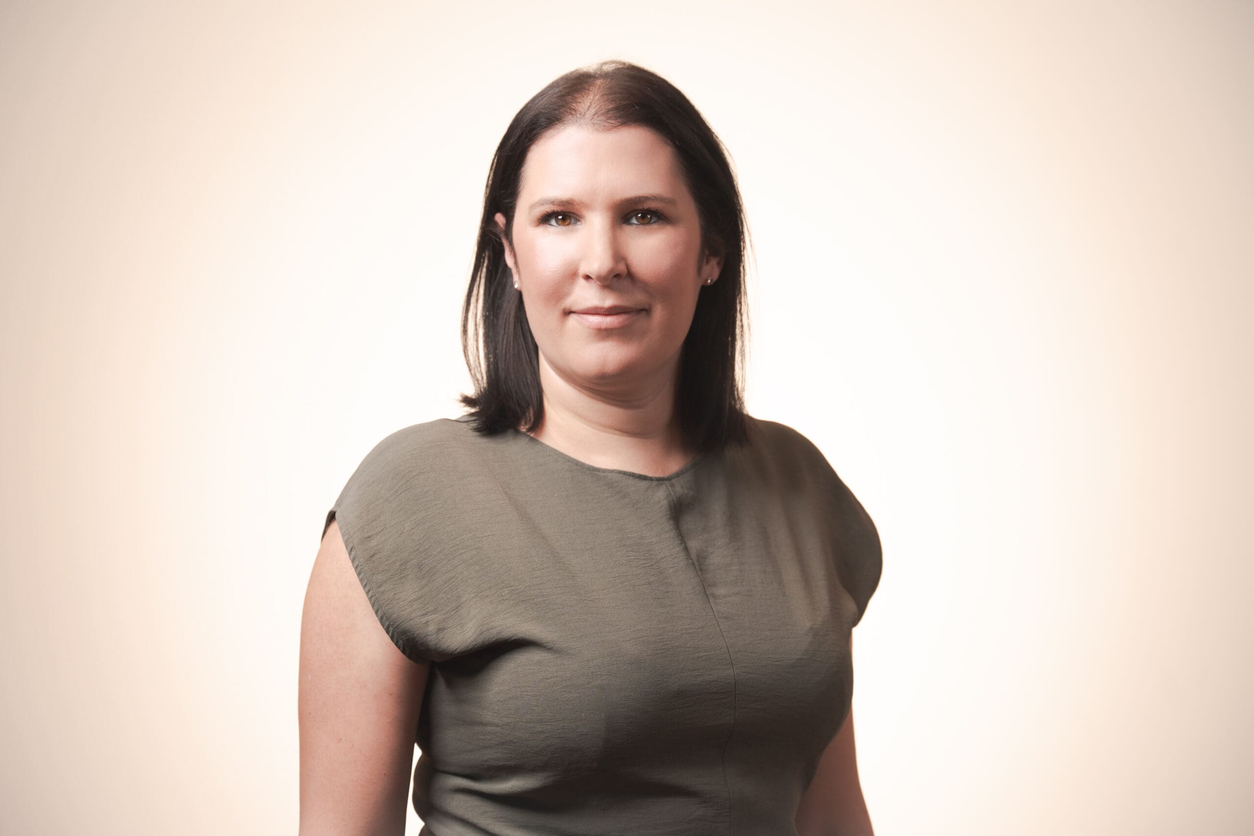 A woman with straight dark hair, wearing a short-sleeved olive green top, stands facing the camera against a light beige background, smiling softly.
